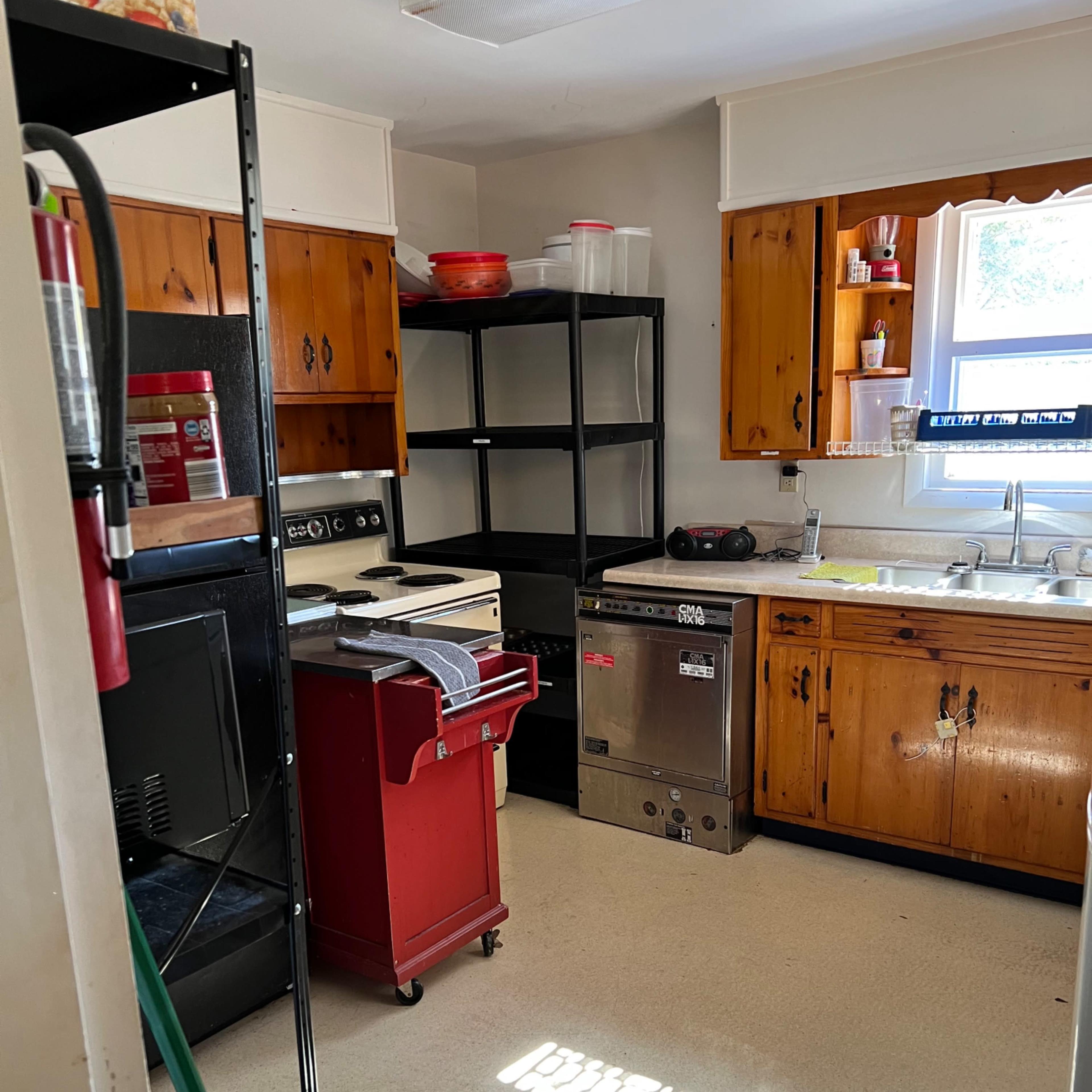 A small kitchen featuring wooden cabinets, a stainless steel dishwasher, an open shelving unit, and various appliances neatly arranged.