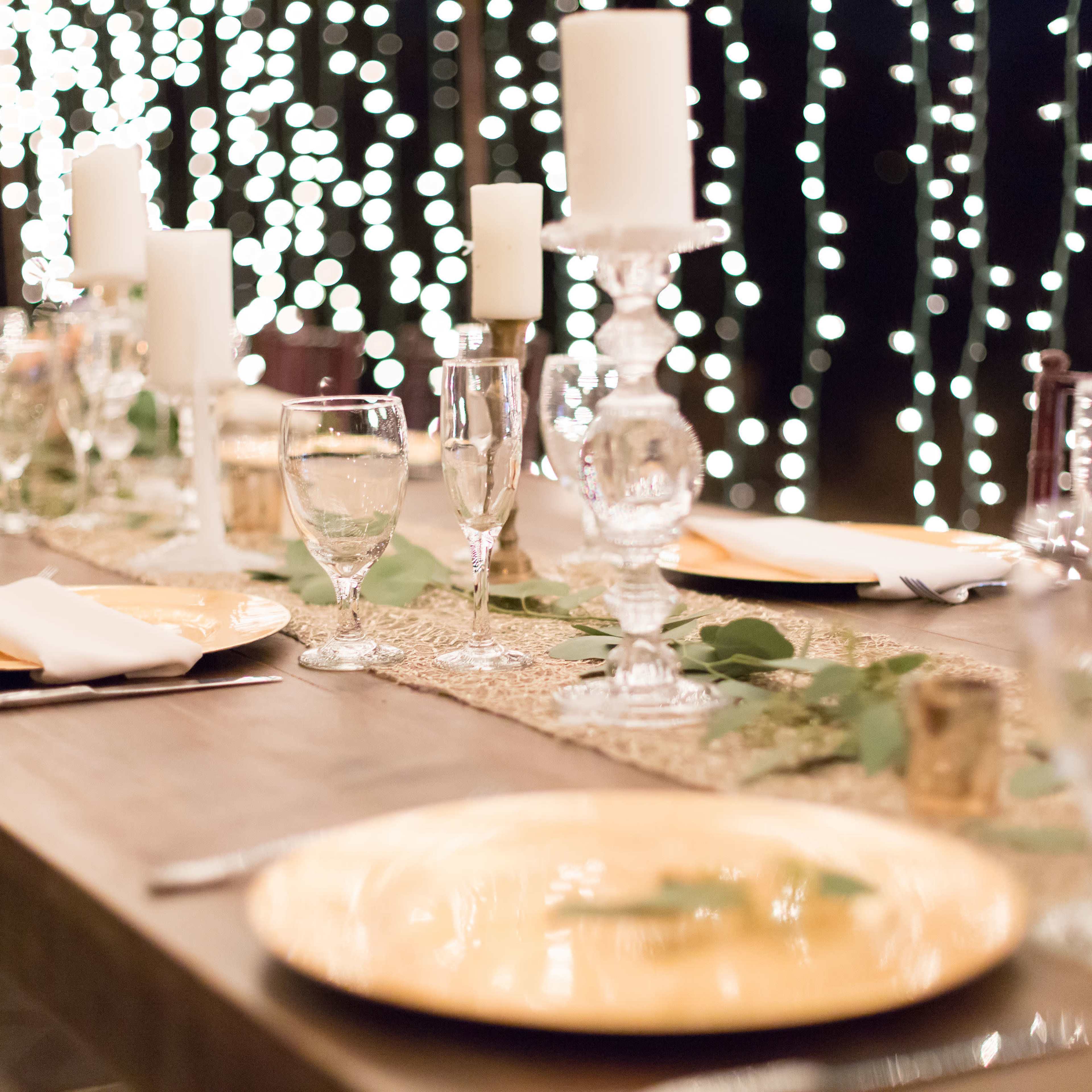 A wooden table is set with glassware and gold plates, adorned with greenery and surrounded by softly glowing string lights.