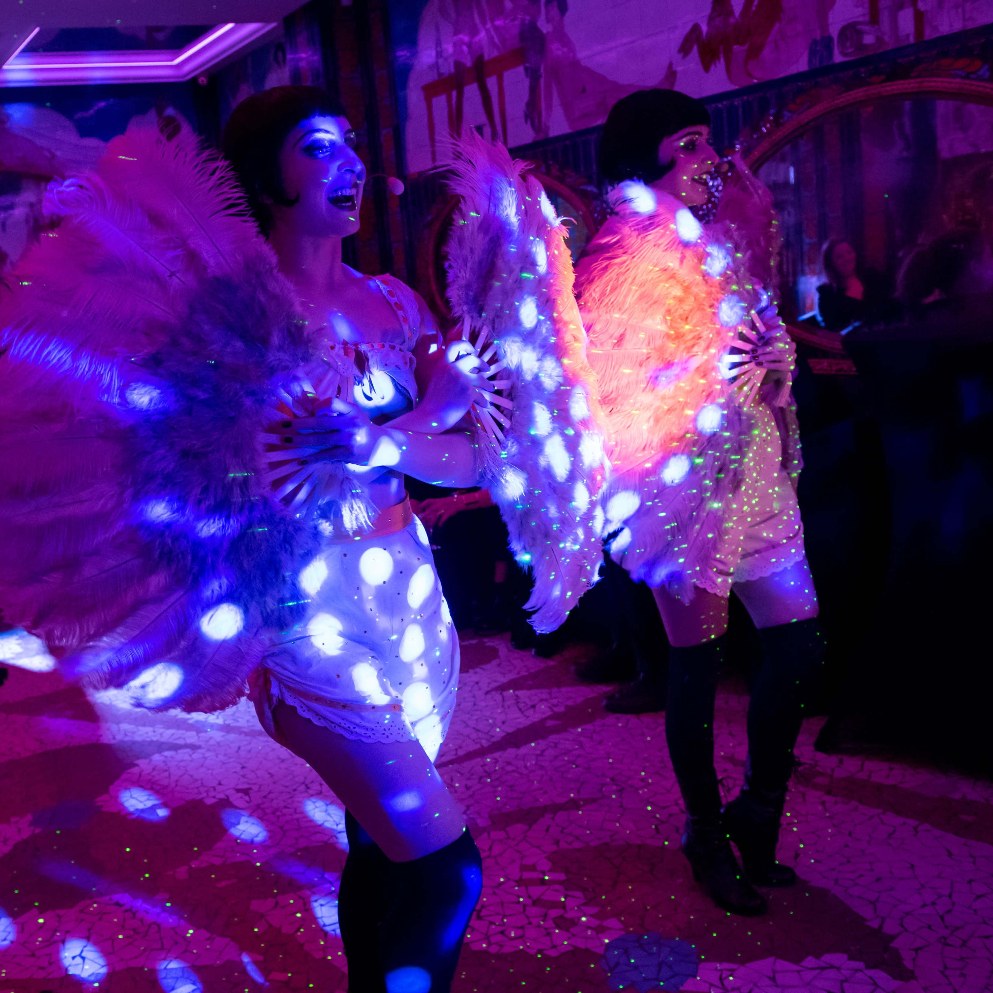 Two performers in feathered costumes with illuminated accents dance on stage in front of an audience.