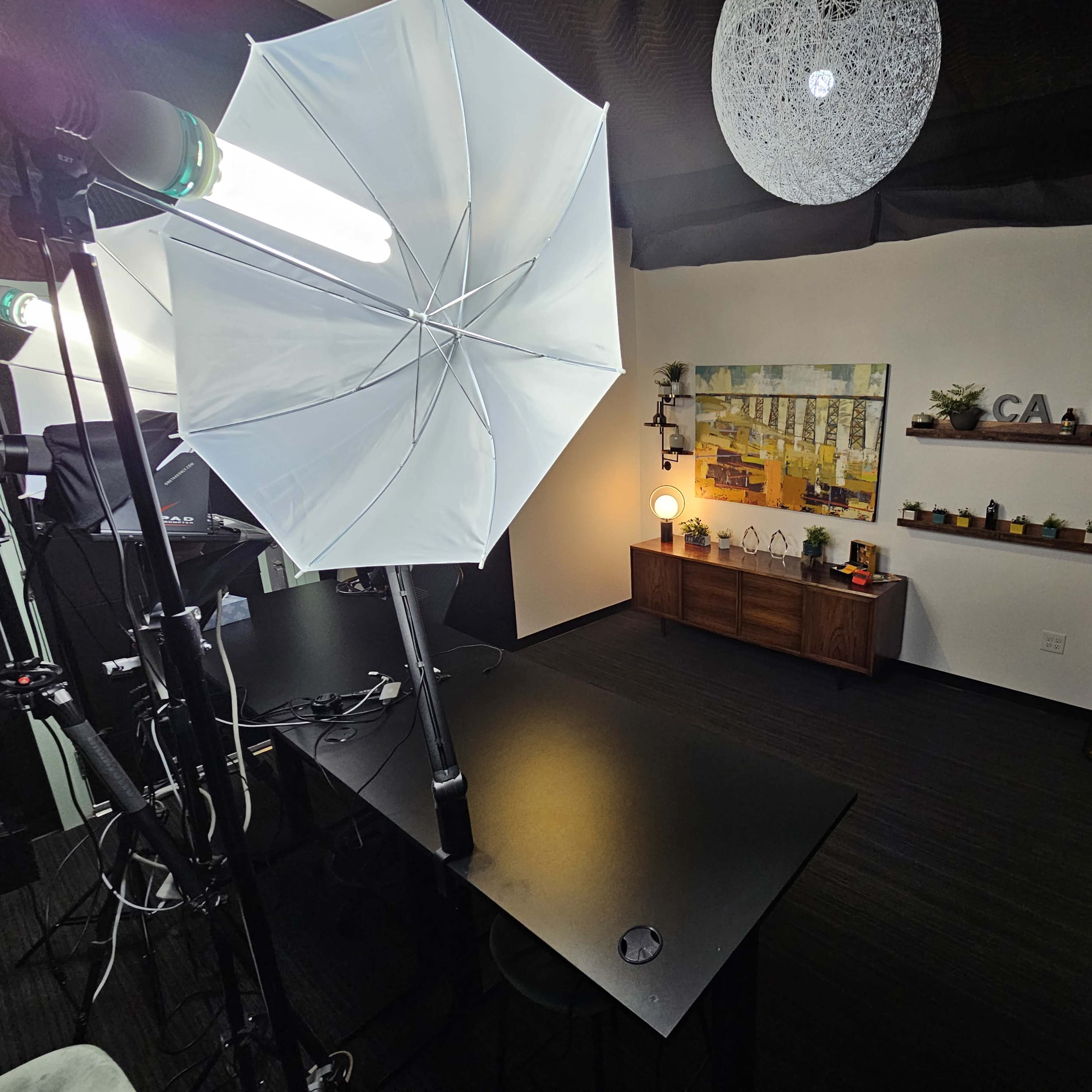 A well-lit studio space with a large umbrella light, a dark table, and decorative shelves on a white wall.