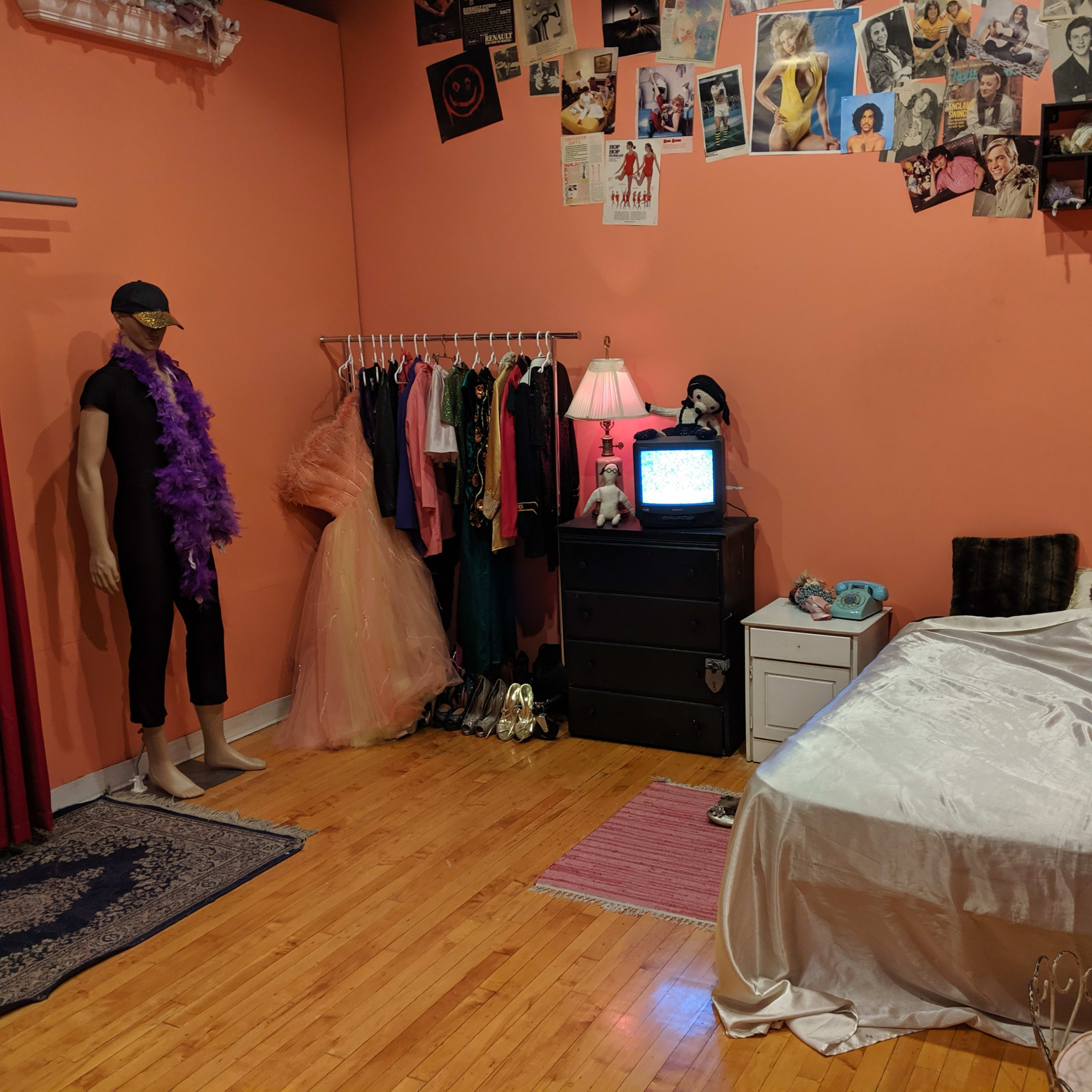 A room with an orange wall, a bed with white bedding, a mannequin dressed in black near a rack of costumes, a dresser, and a vintage television on a table.