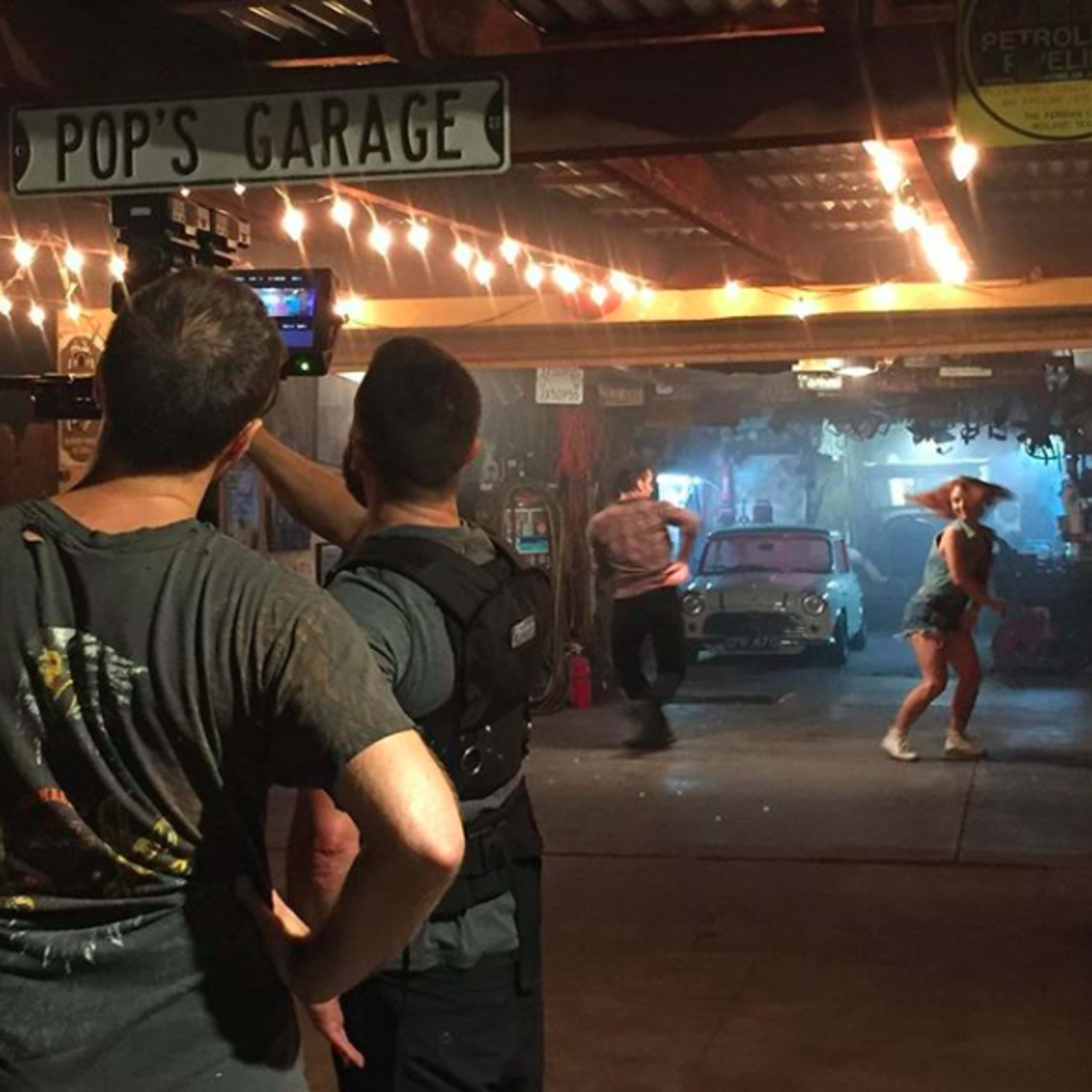 A film crew captures a dance scene inside a garage adorned with string lights and a vintage car in the background.