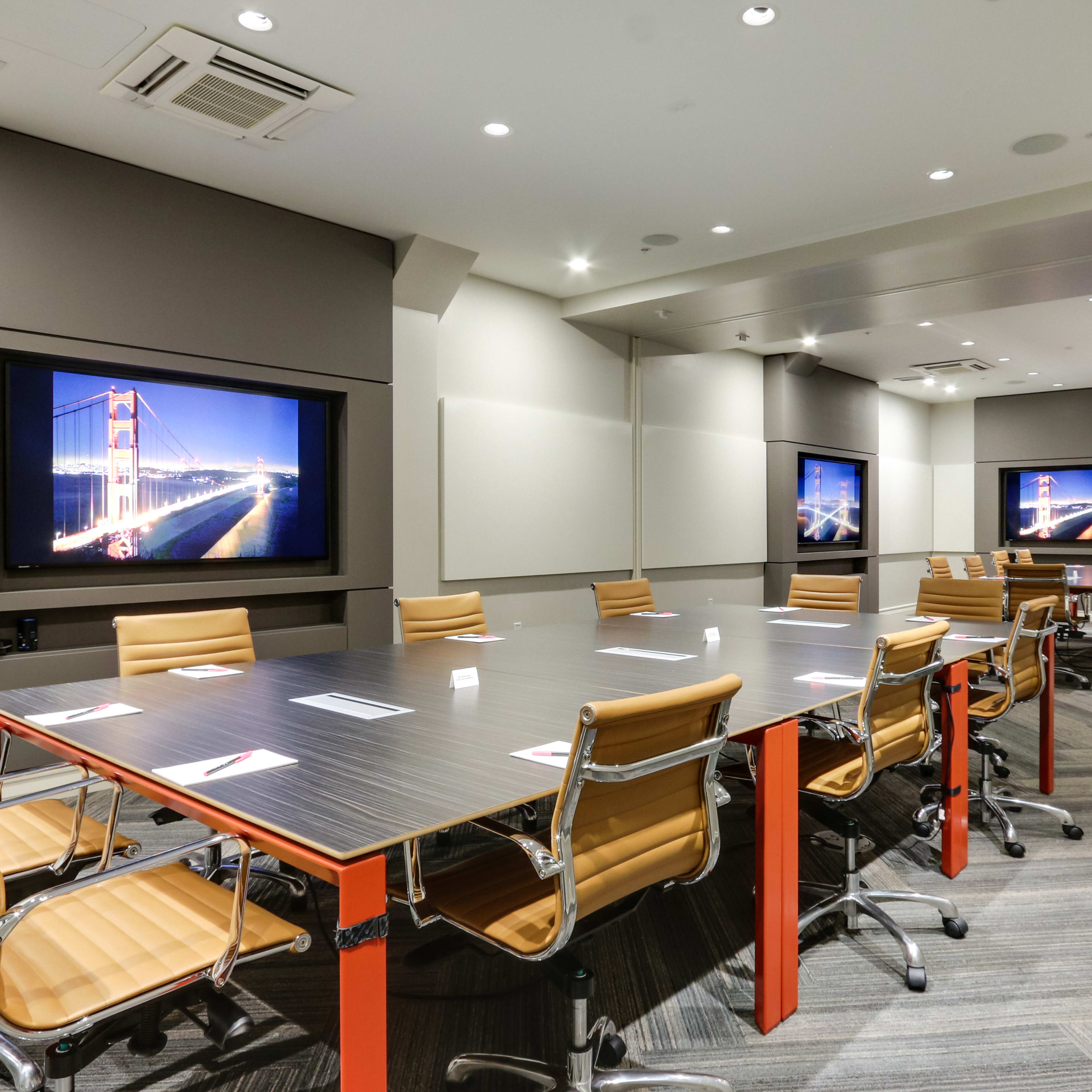 A modern conference room equipped with a large table, ergonomic chairs, and two wall-mounted screens displaying an image of the Golden Gate Bridge.