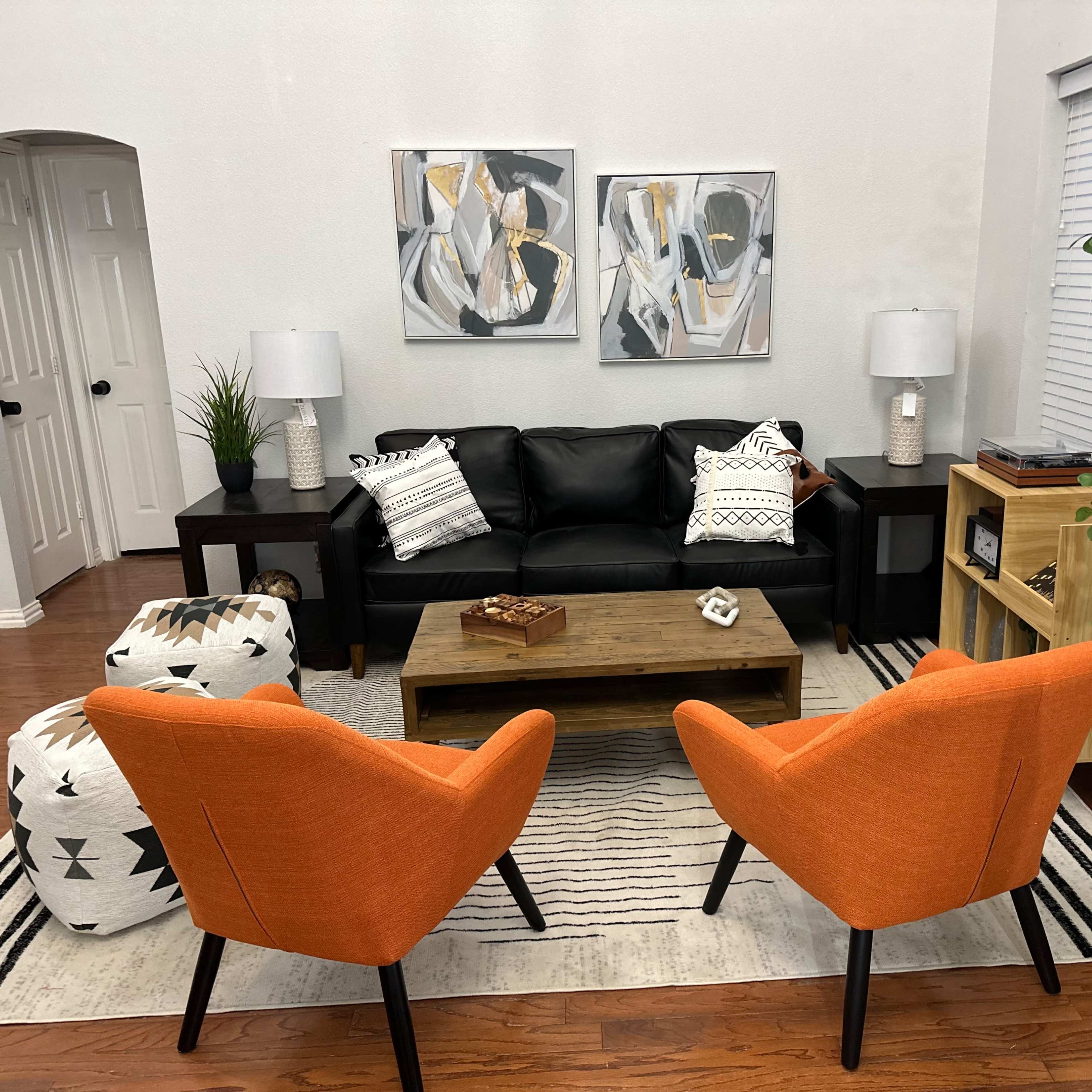 A modern living room features a black leather sofa, two orange chairs, a wooden coffee table, and abstract artwork on the walls.