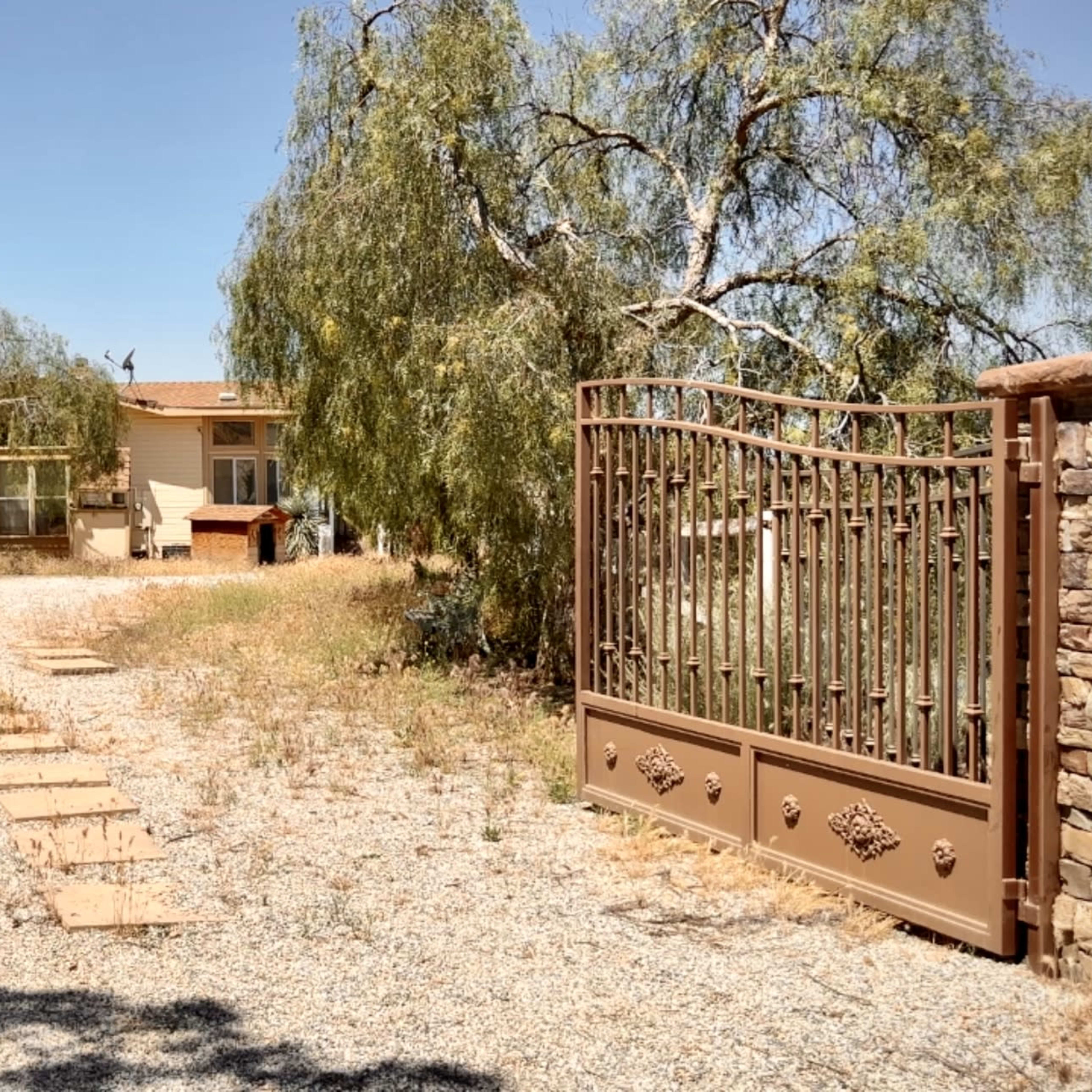 A gated entrance leads to a house surrounded by trees and gravel on a sunny day.