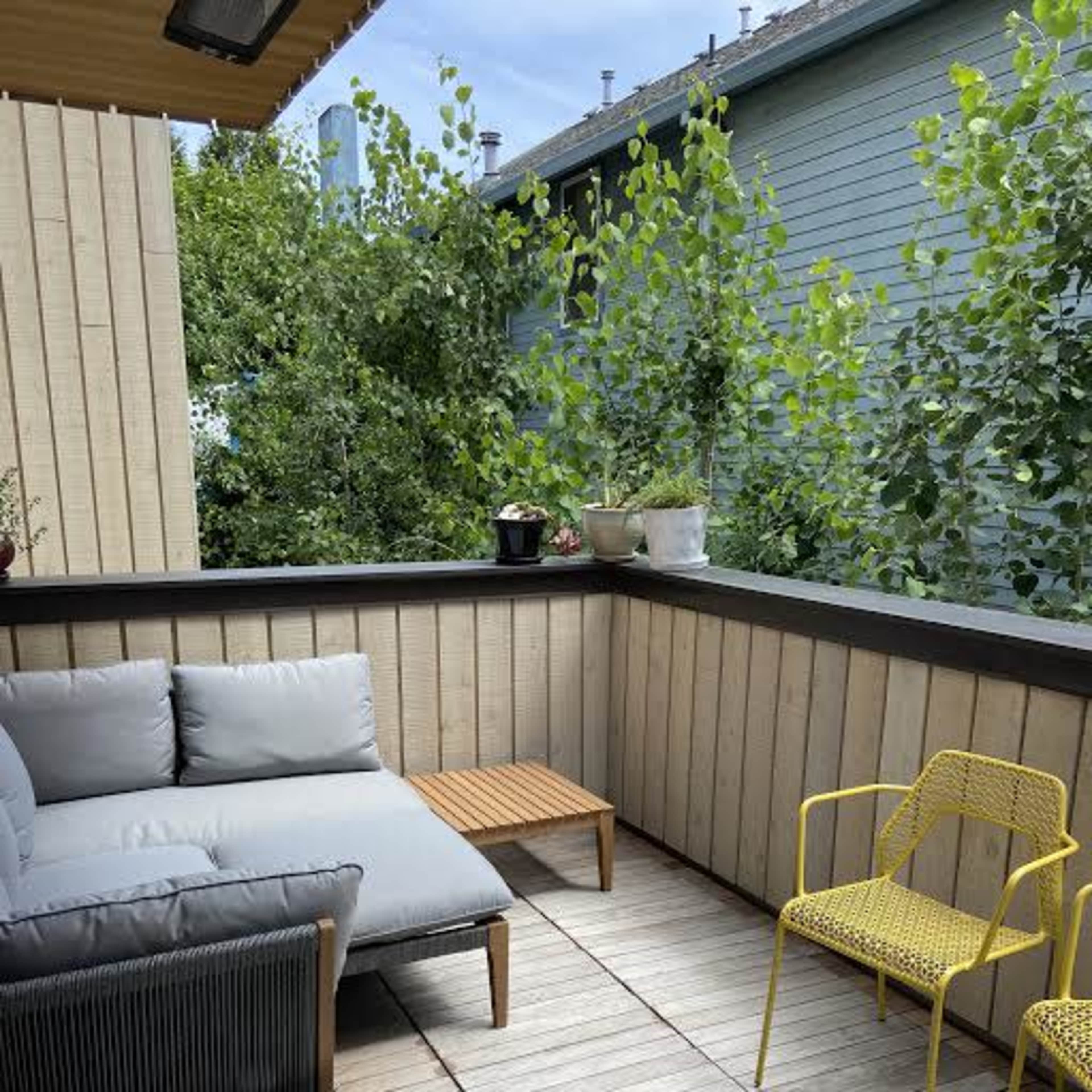 A cozy balcony features a gray sofa, a wooden coffee table, and two yellow chairs surrounded by greenery.