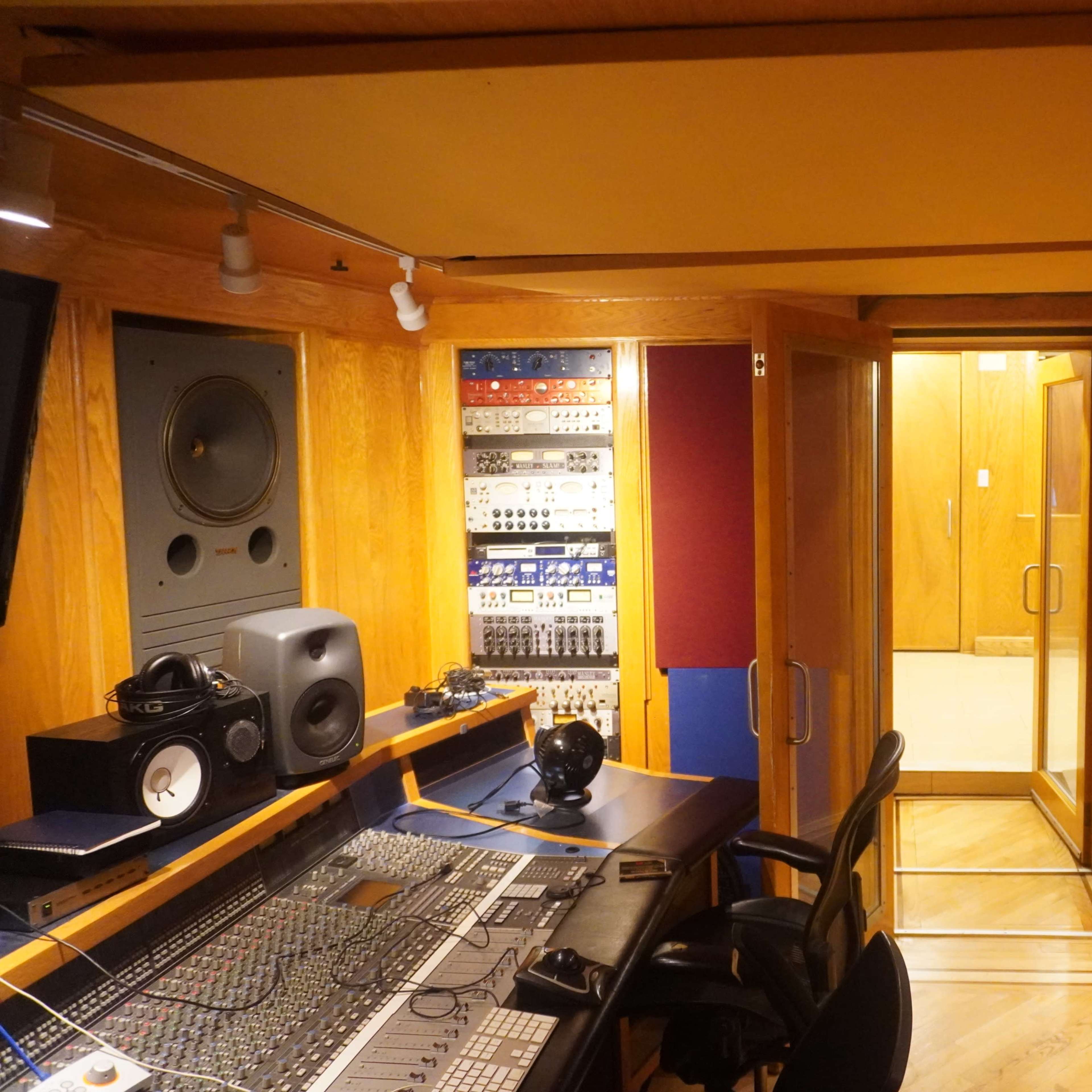 A sound studio featuring a control desk with audio equipment, a red couch, and a glass door leading to an adjacent room.