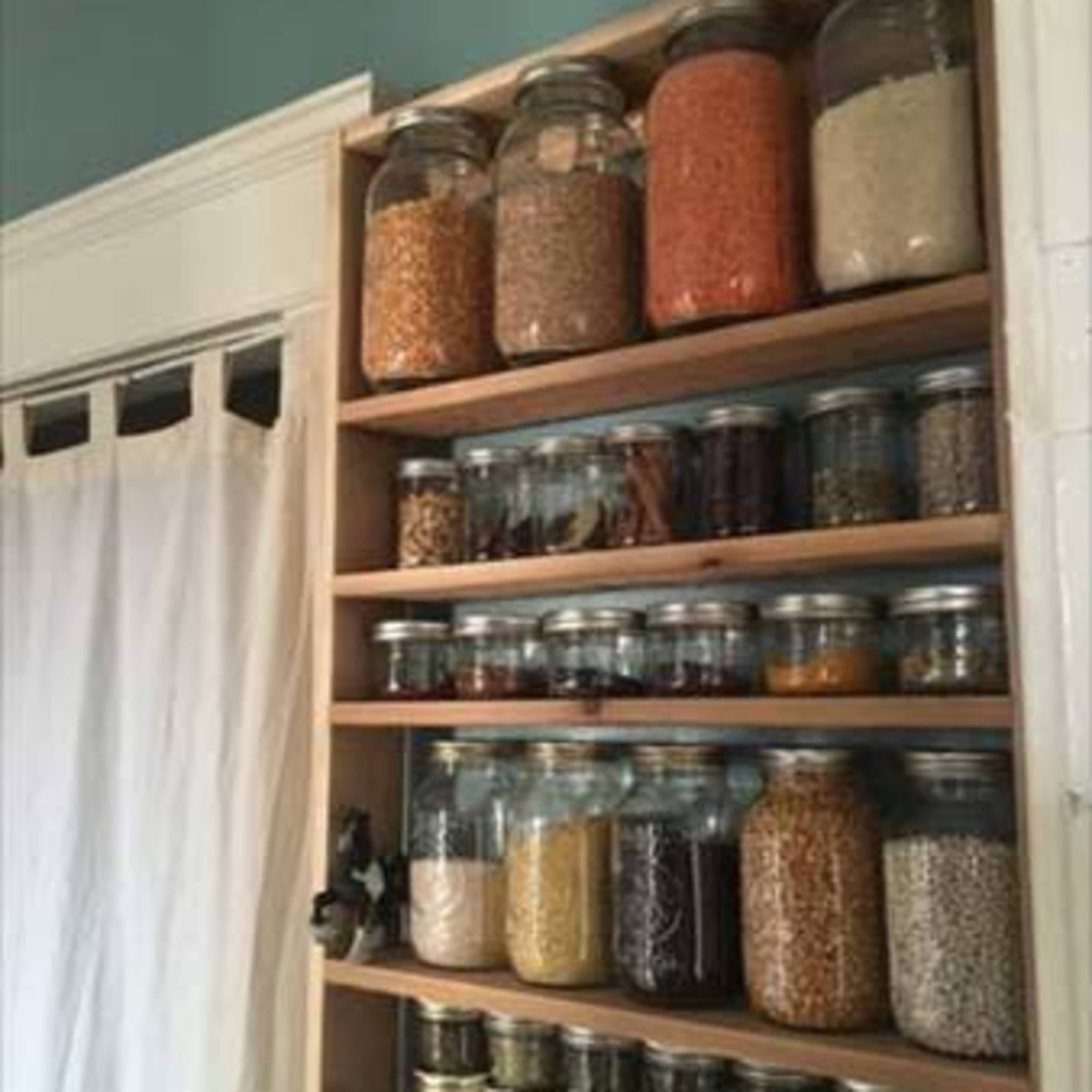 A wooden shelf filled with various glass jars containing colorful grains and spices, organized neatly against a muted wall.