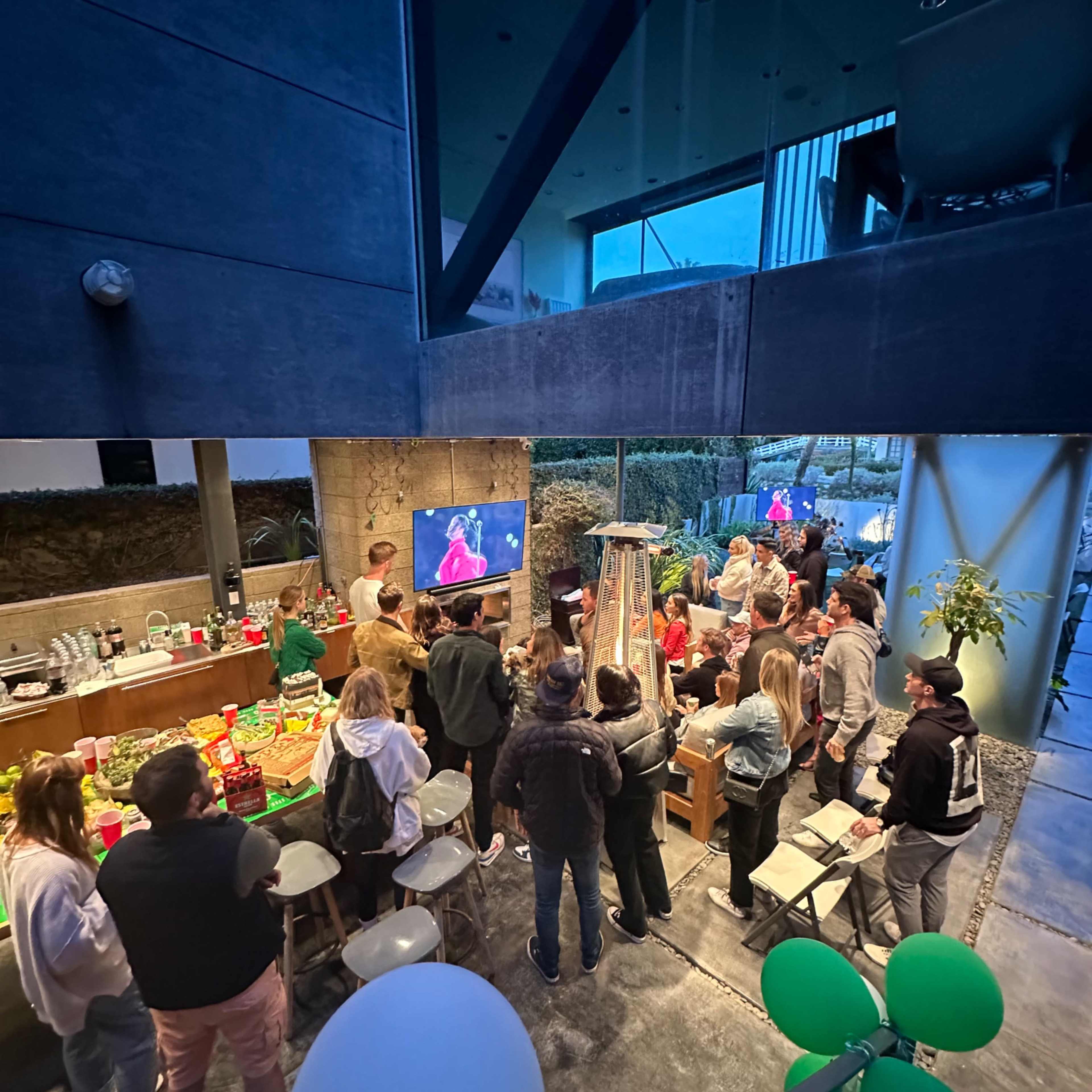 A group of people gathers outdoors around a large television, enjoying food and drinks in a modern setting.