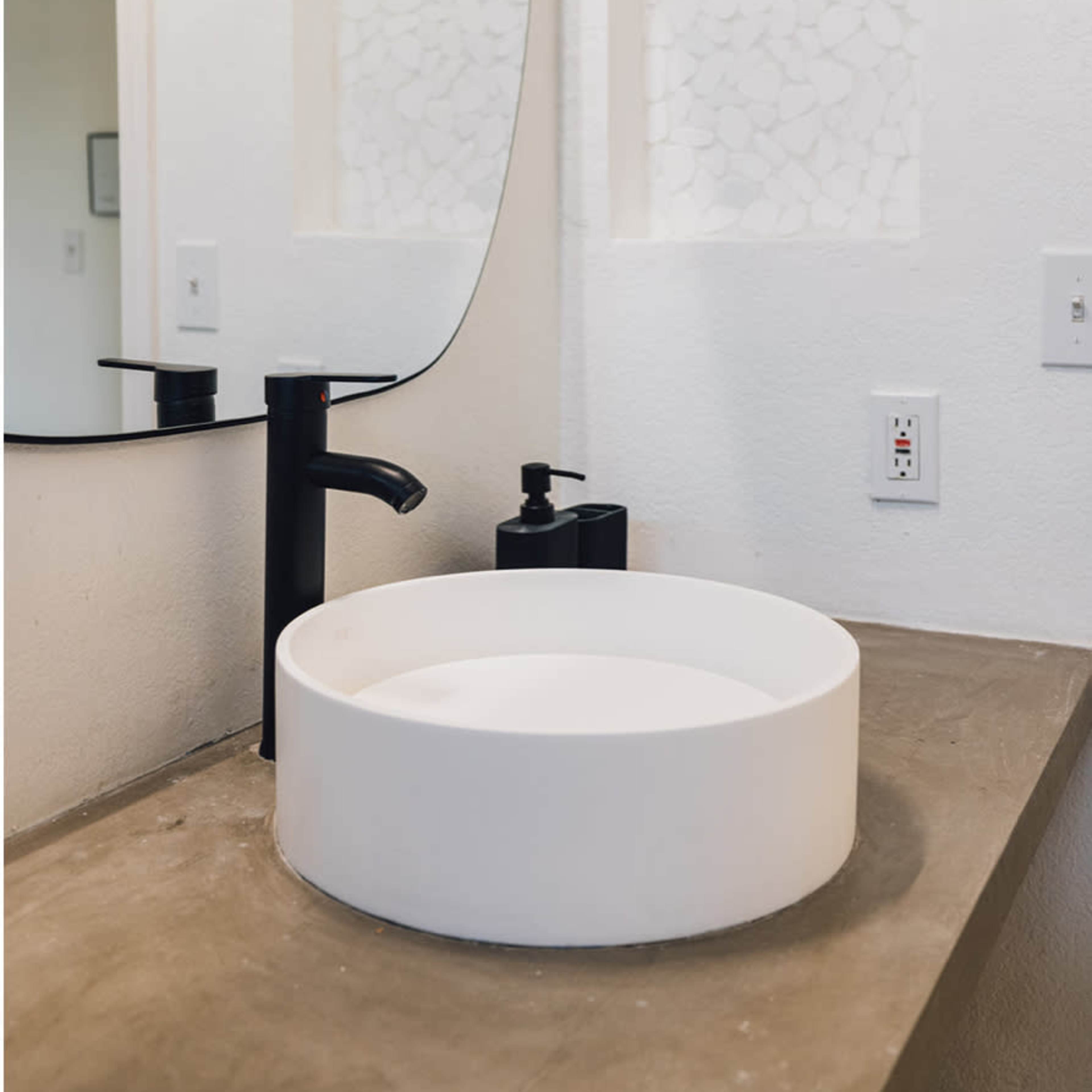 A modern bathroom sink made of white circular porcelain positioned on a concrete countertop, with a sleek black faucet and a curved mirror above it.