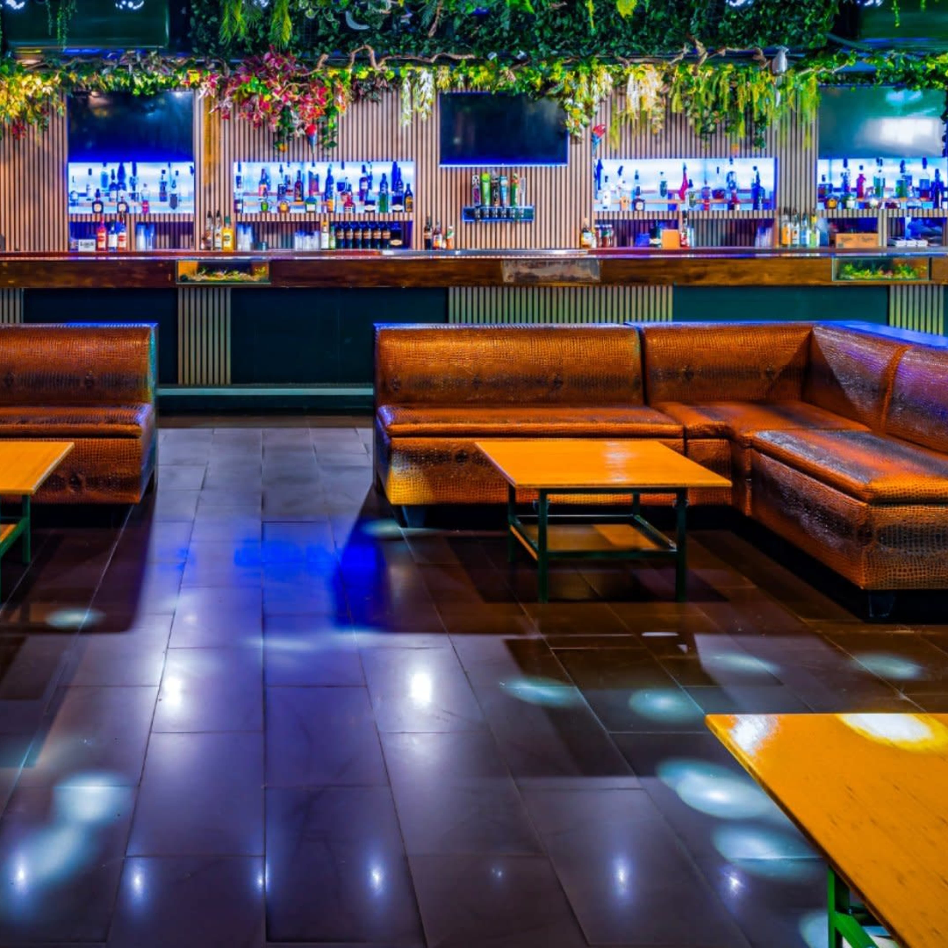 A bar area with brown leather couches and wooden tables, illuminated by colored lights and adorned with greenery.