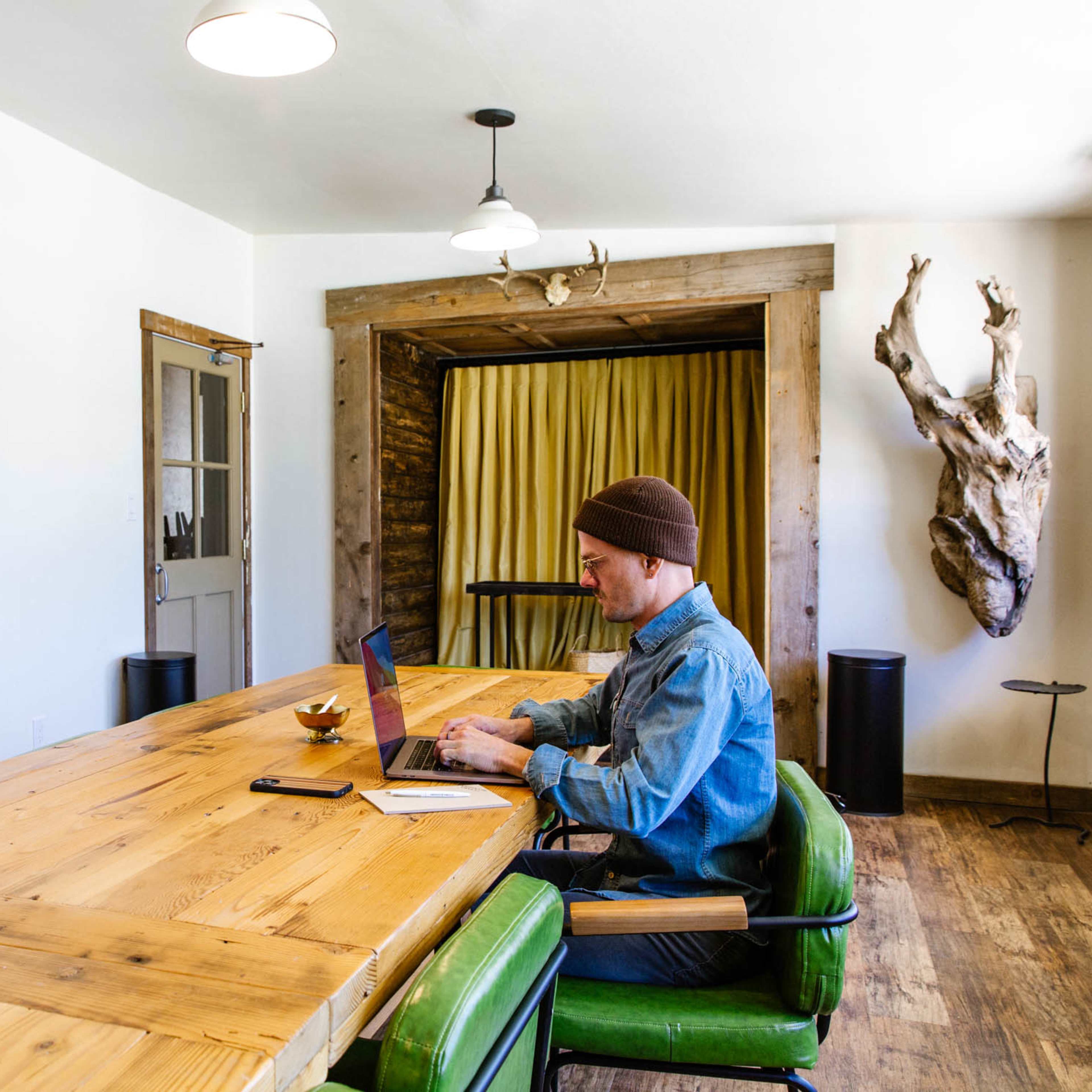 A person works on a laptop at a large wooden table in a bright, minimalist room with wooden accents and a curtain in the background.