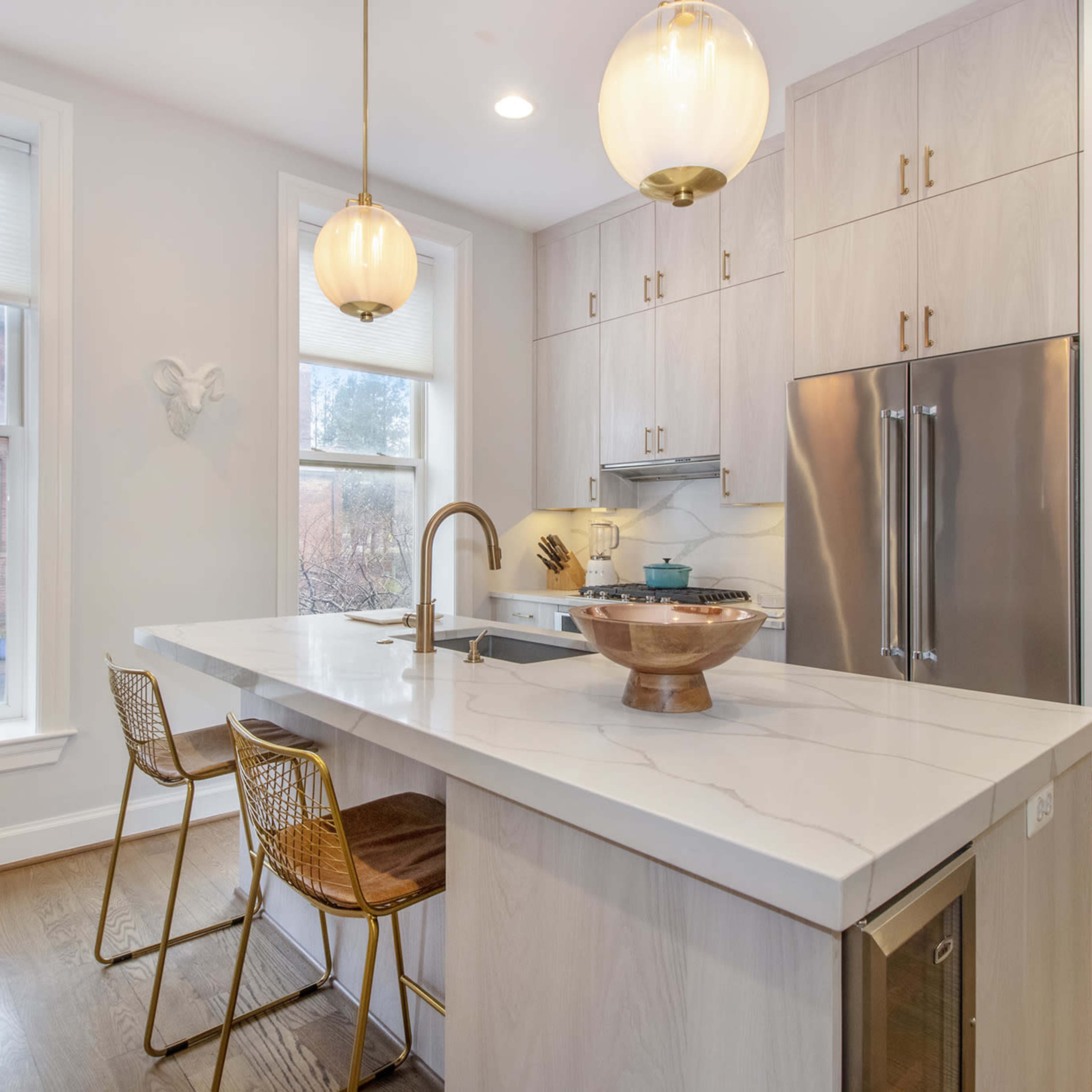 A modern kitchen with a large white island, two gold-accented bar stools, and pendant lighting above, featuring wooden cabinetry and stainless steel appliances.