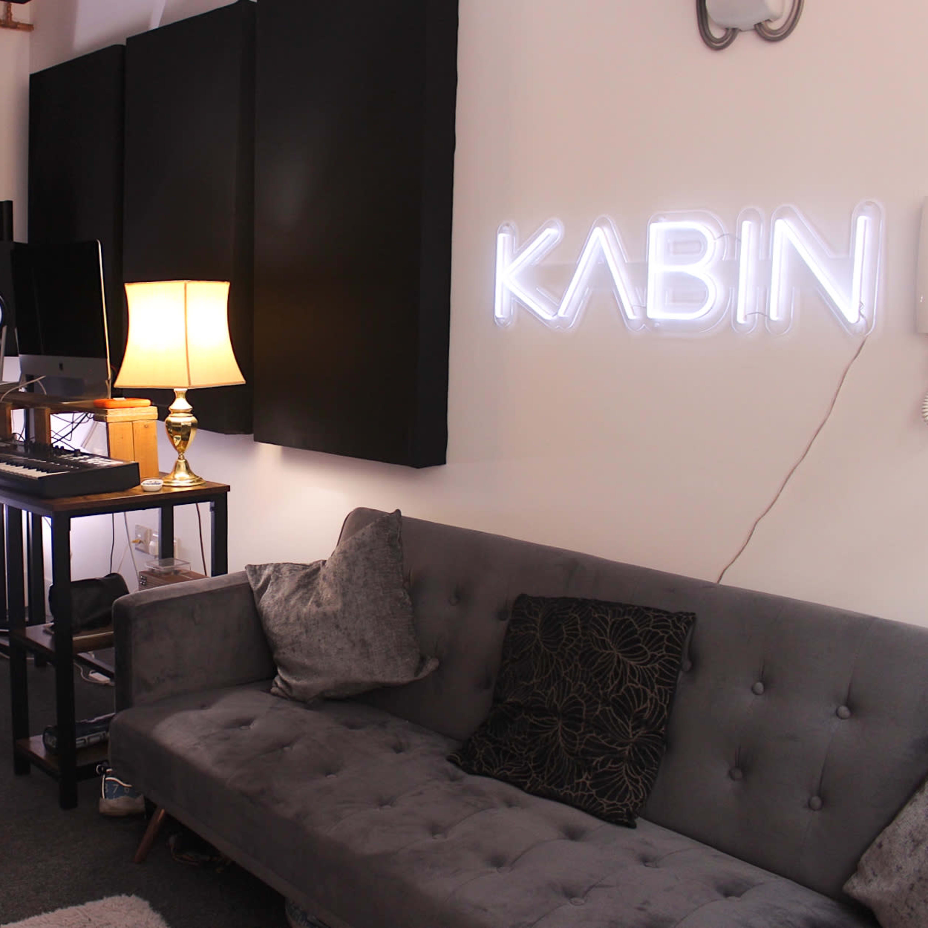 A music studio featuring a gray sofa, a keyboard on a stand, a computer monitor, and soundproofing panels on the walls.