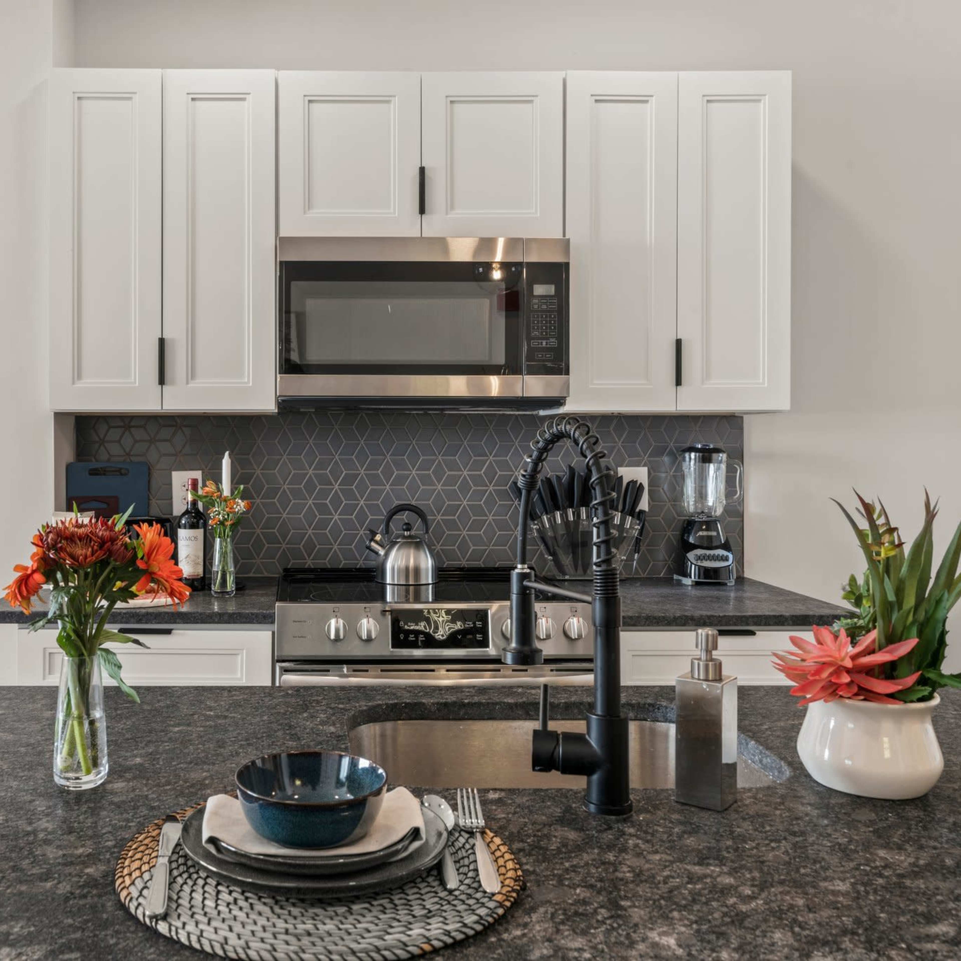 A modern kitchen features a countertop with decorative bowls, flowers in vases, and stainless steel appliances against a geometric-tiled backsplash.