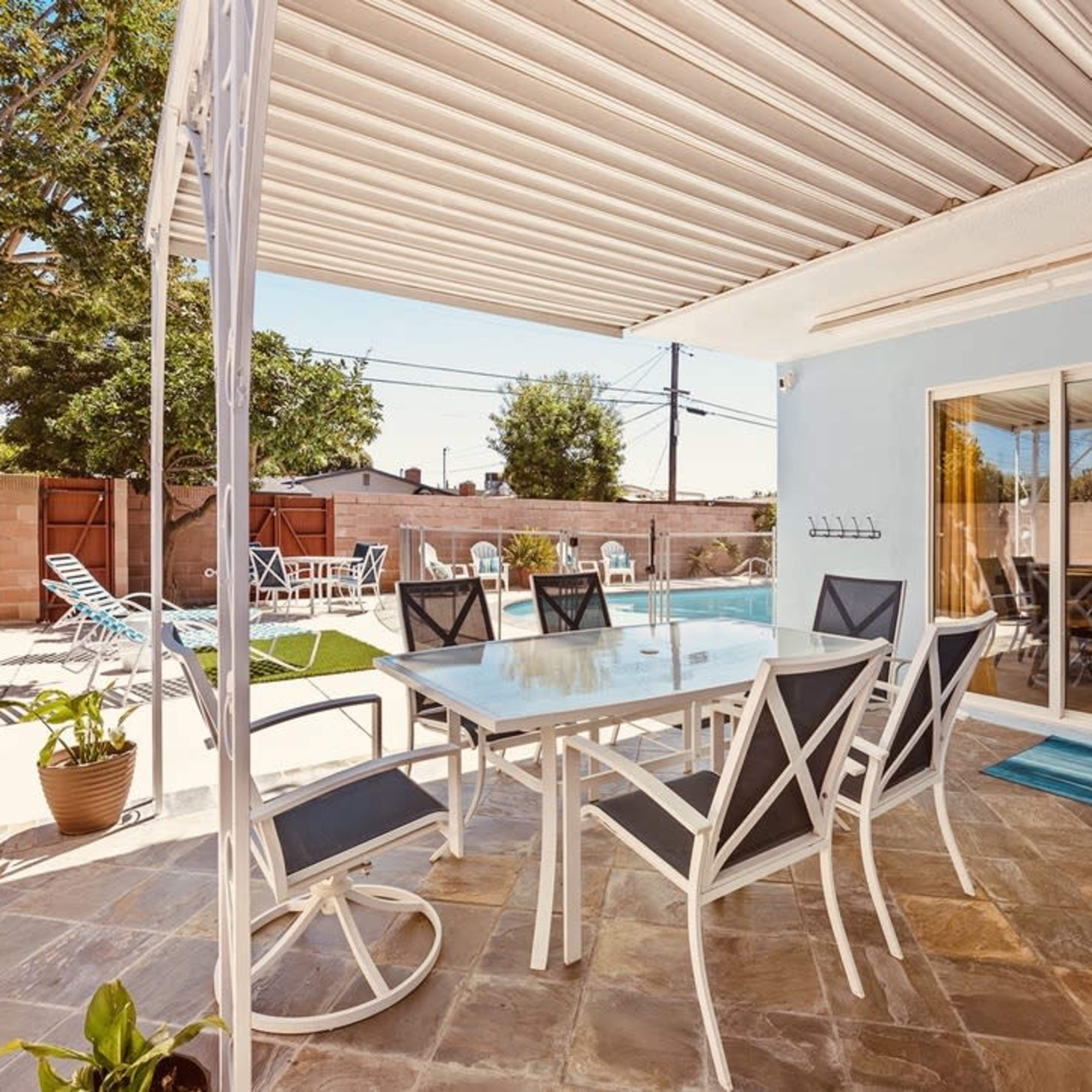 A patio area features a dining table surrounded by chairs, adjacent to a pool, with lawn chairs in the background under a shaded awning.