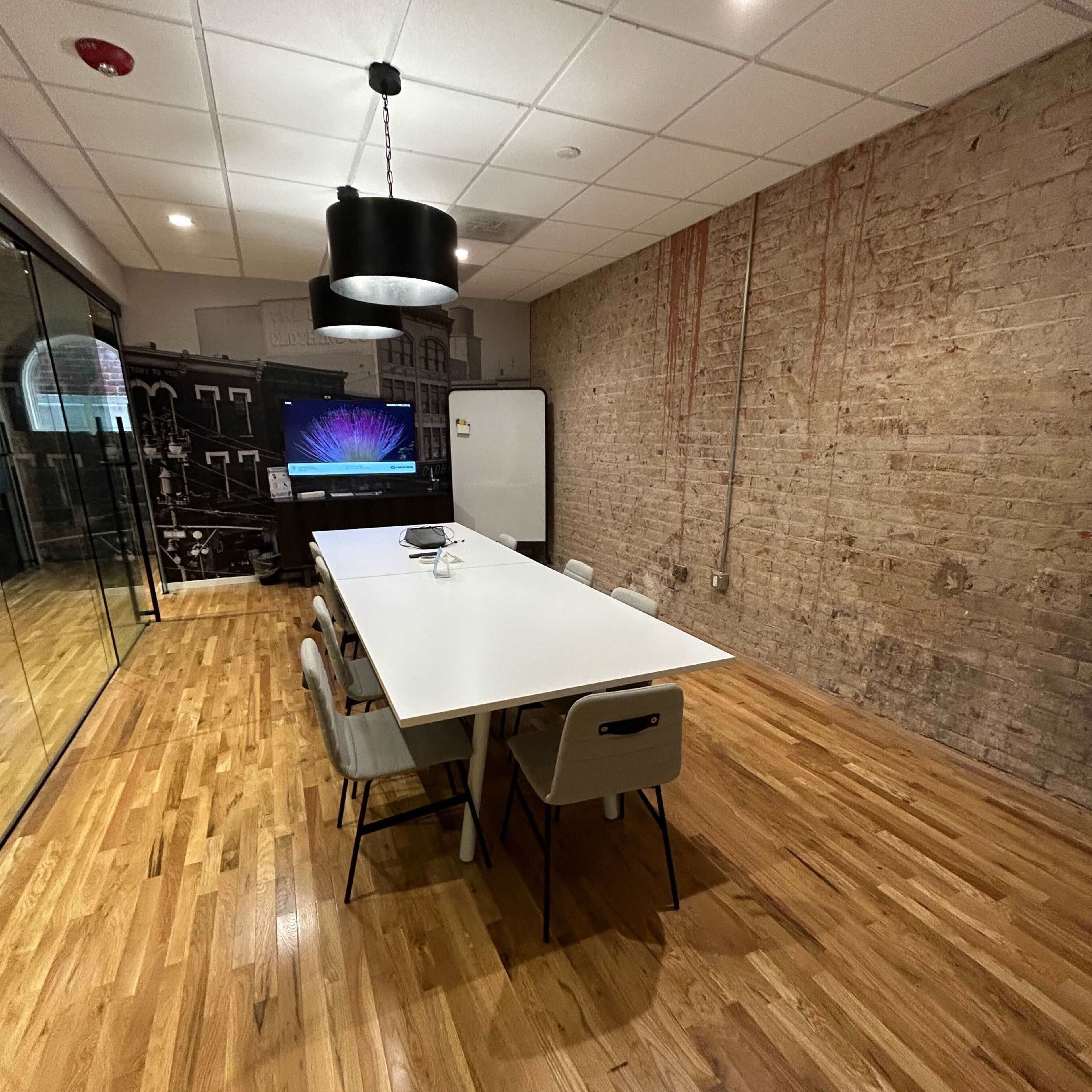 A modern conference room features a long white table surrounded by chairs, with a large screen on one wall and a brick accent wall.