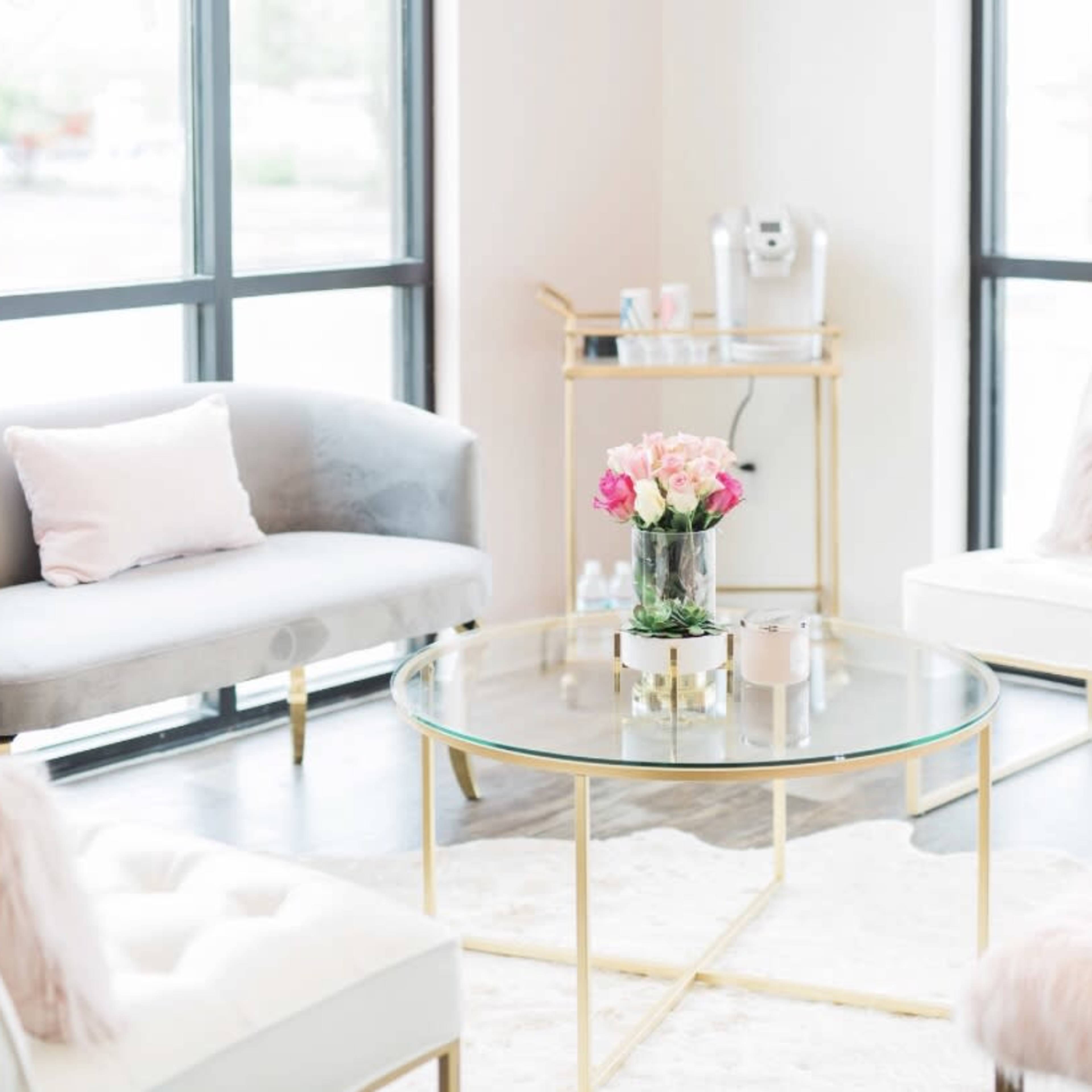 A modern waiting room featuring a glass coffee table surrounded by a gray couch and two white chairs, with a bouquet of flowers on the table.