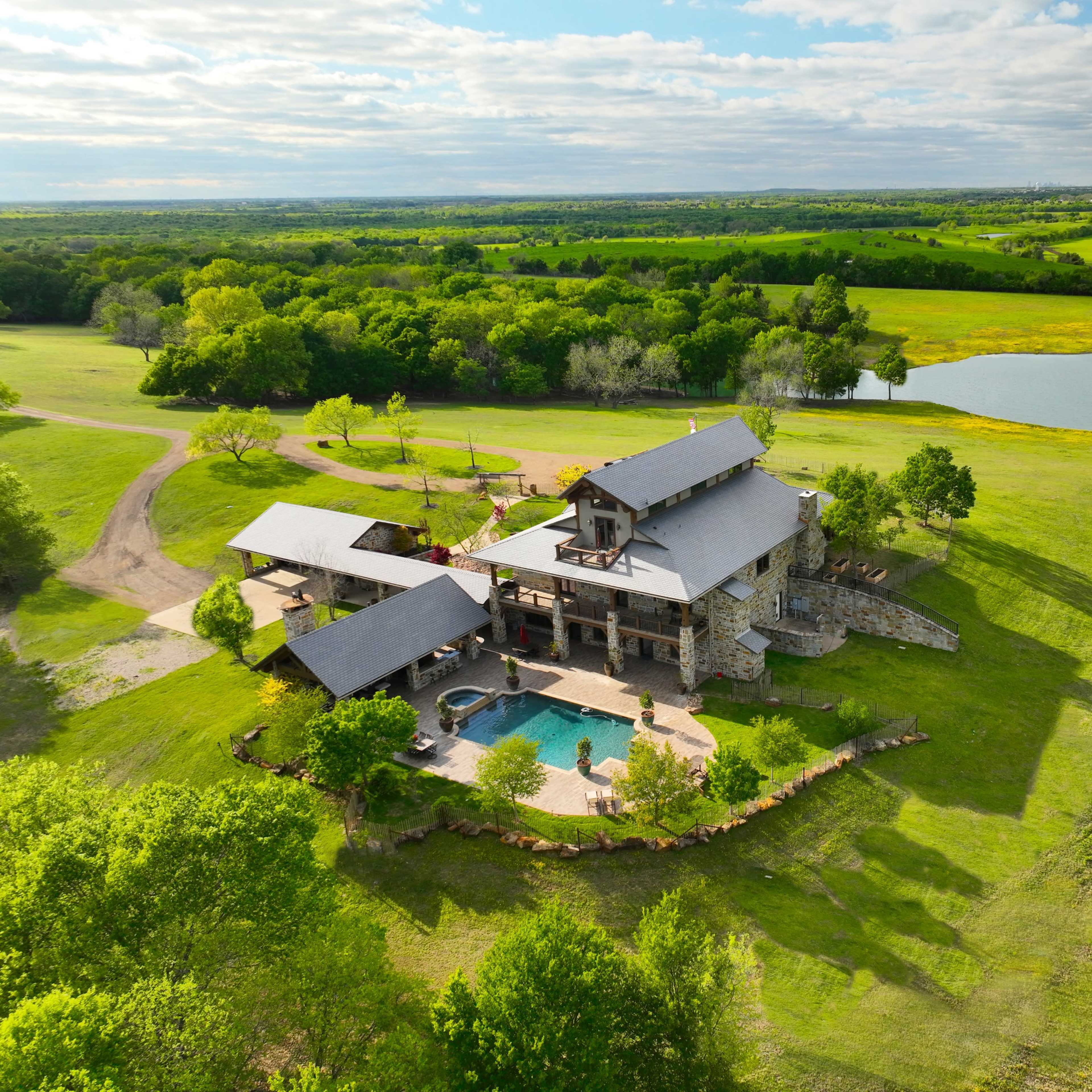 A large, modern house with a swimming pool, surrounded by greenery and a pond in a rural landscape.
