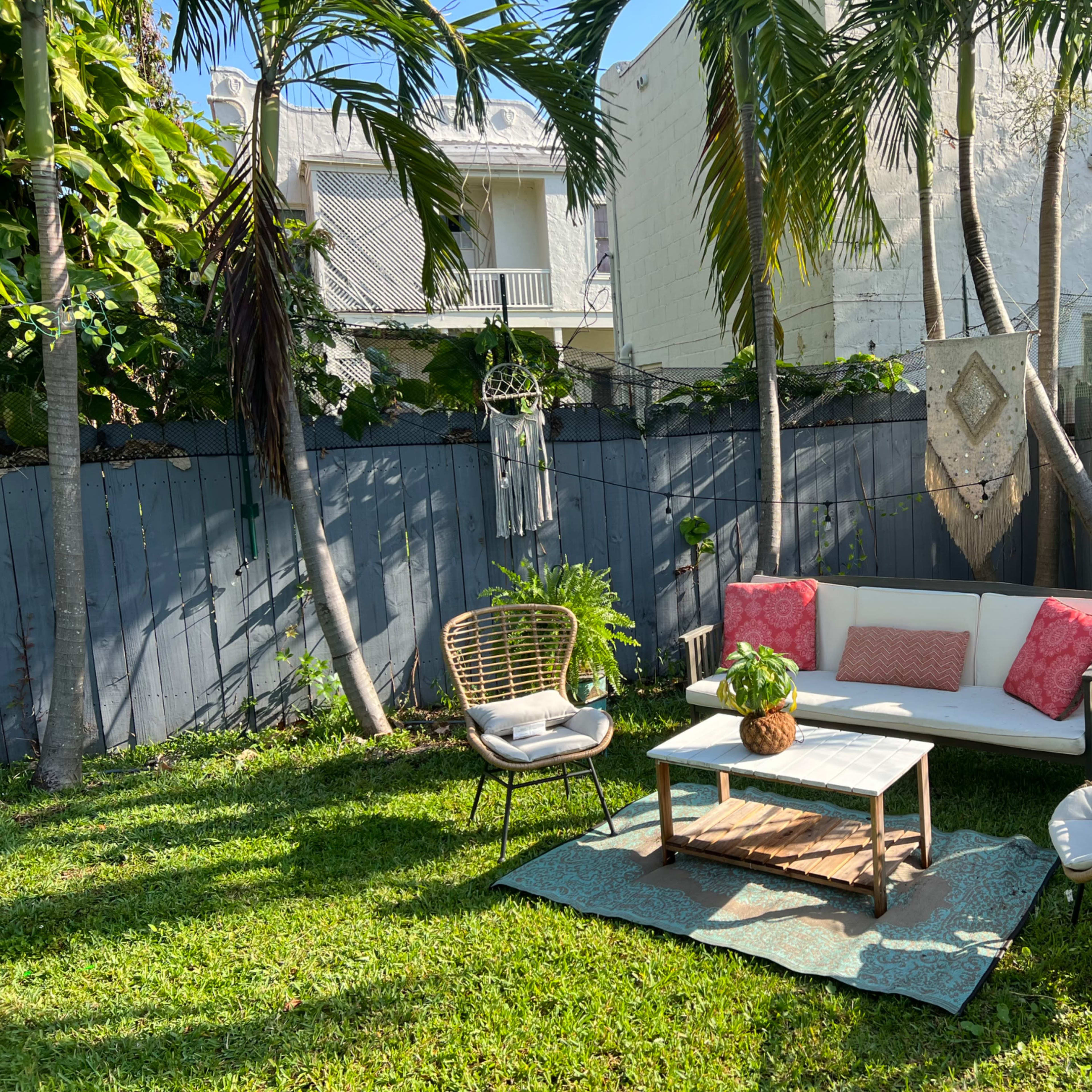 A small outdoor seating area features a white couch with pink cushions, a wooden coffee table, and three chairs surrounded by lush greenery and a wooden fence.