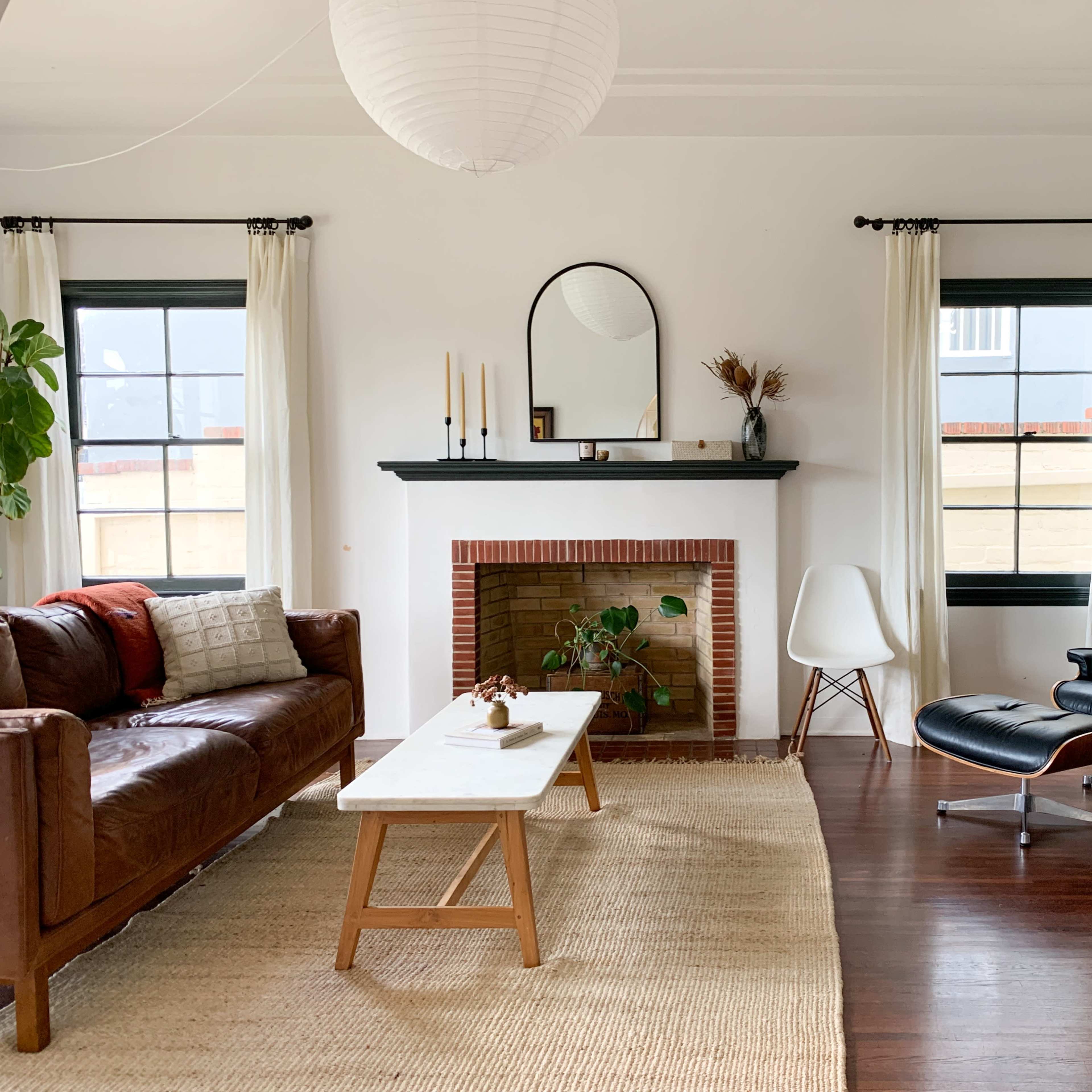 A living room featuring a brown leather sofa, a white coffee table, a fireplace with a mounted mirror, and a cozy arrangement of plants, guitars, and a plush chair.