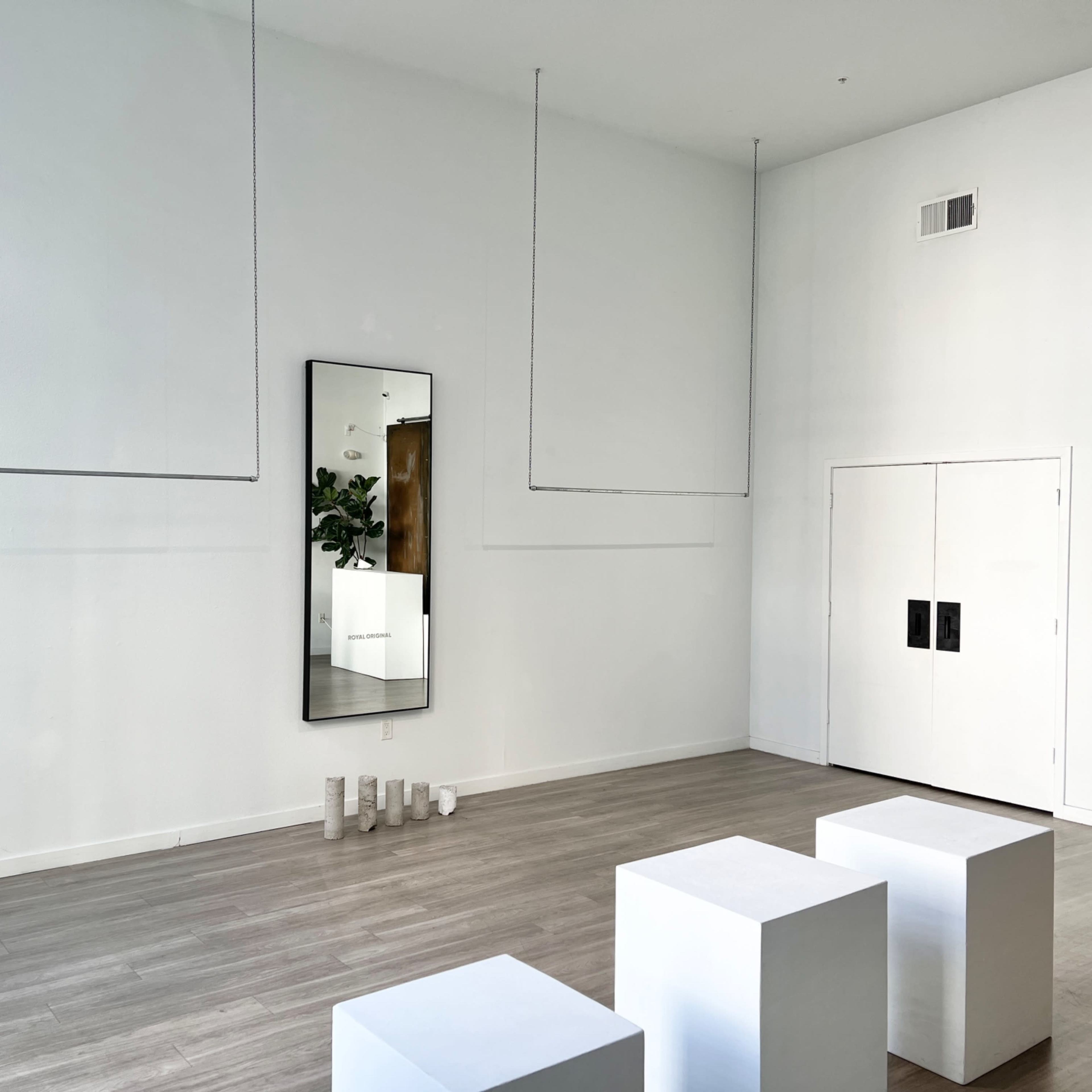 A minimalist interior space with white walls, hanging elements, a large mirror reflecting the room, three white cubes, and a closed double door.