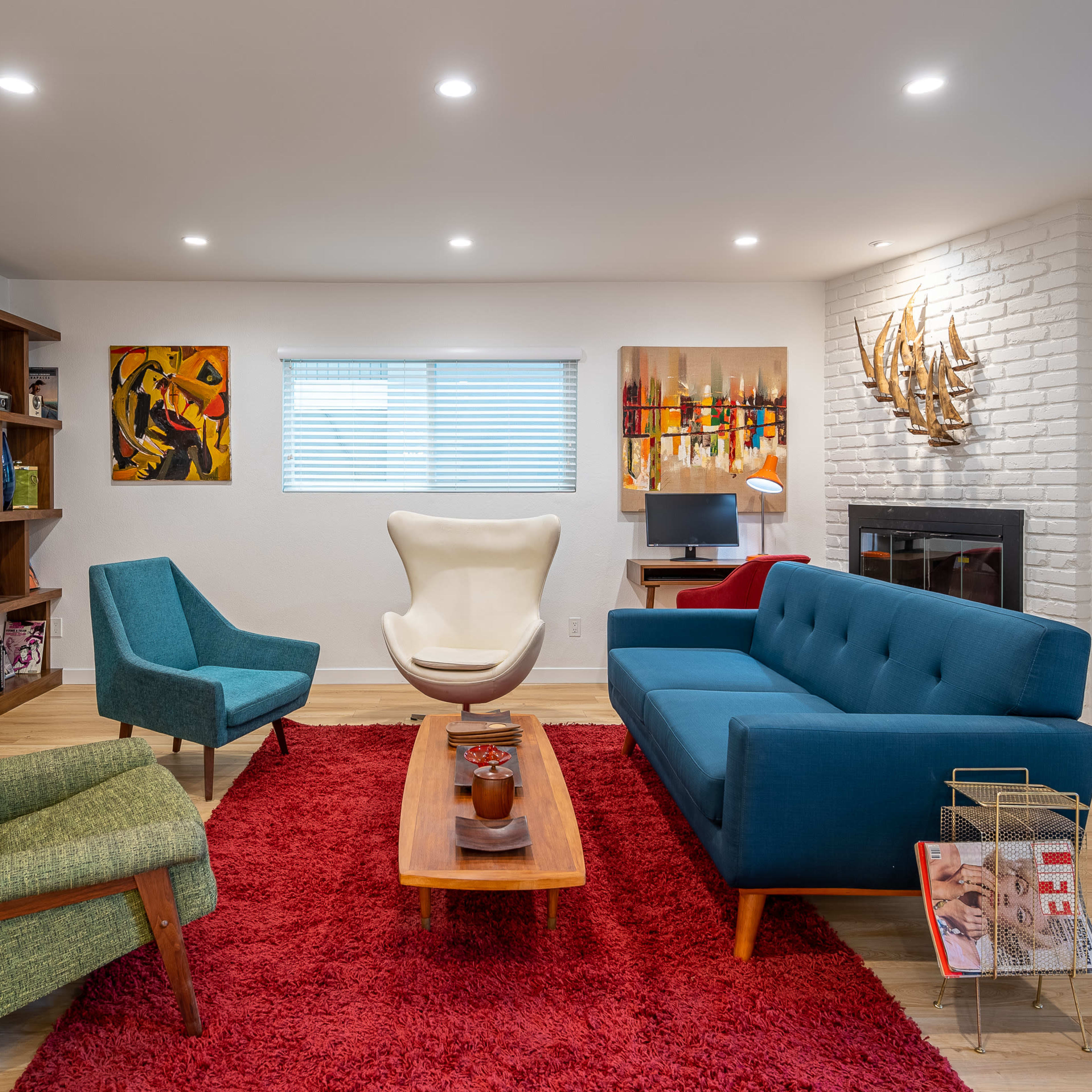 A modern living room featuring a mixture of teal and green seating, a wooden coffee table, a red rug, and shelves with decorative items against a white brick wall.