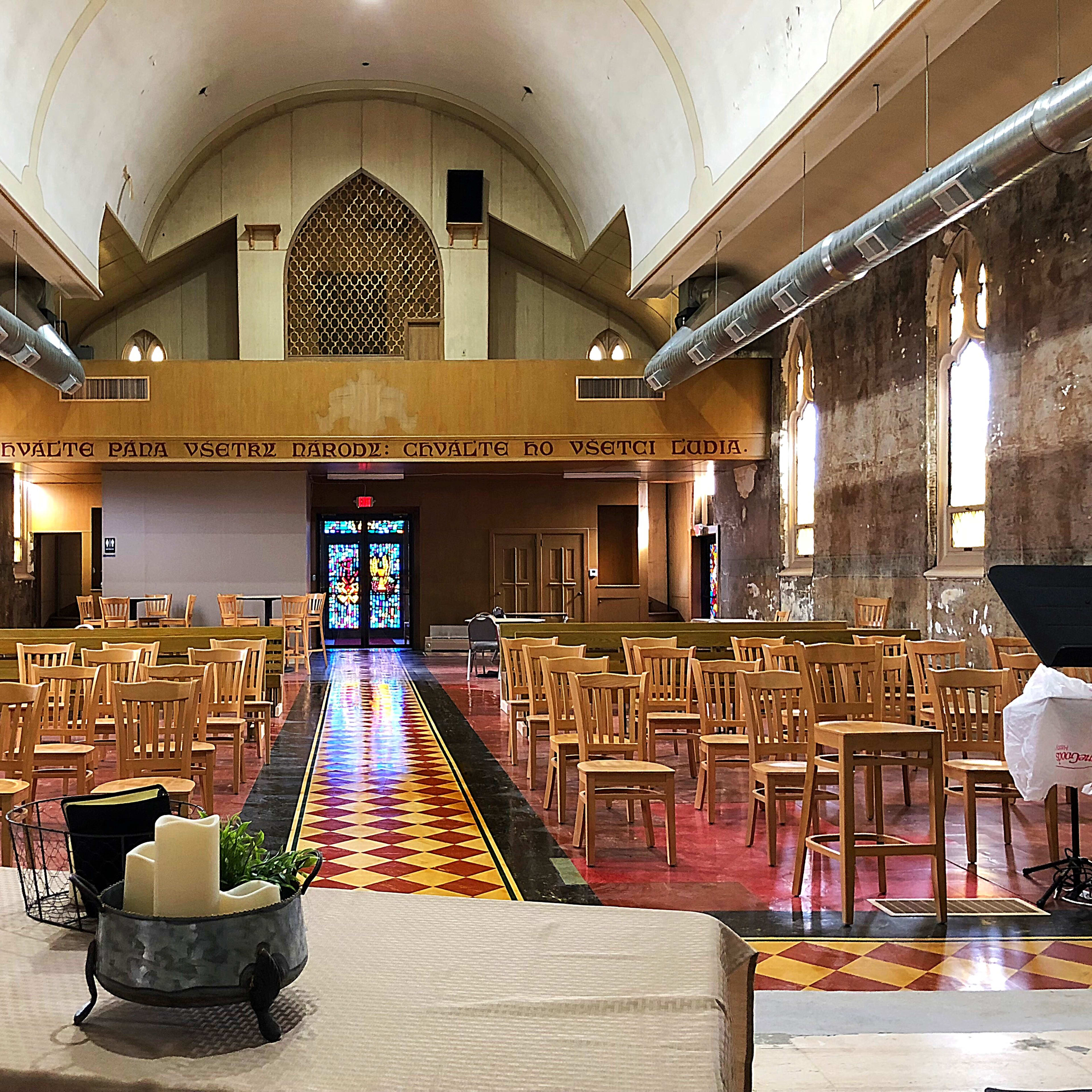 The interior of a large, empty church with wooden chairs arranged in rows and a colorful stained glass window at the far end.