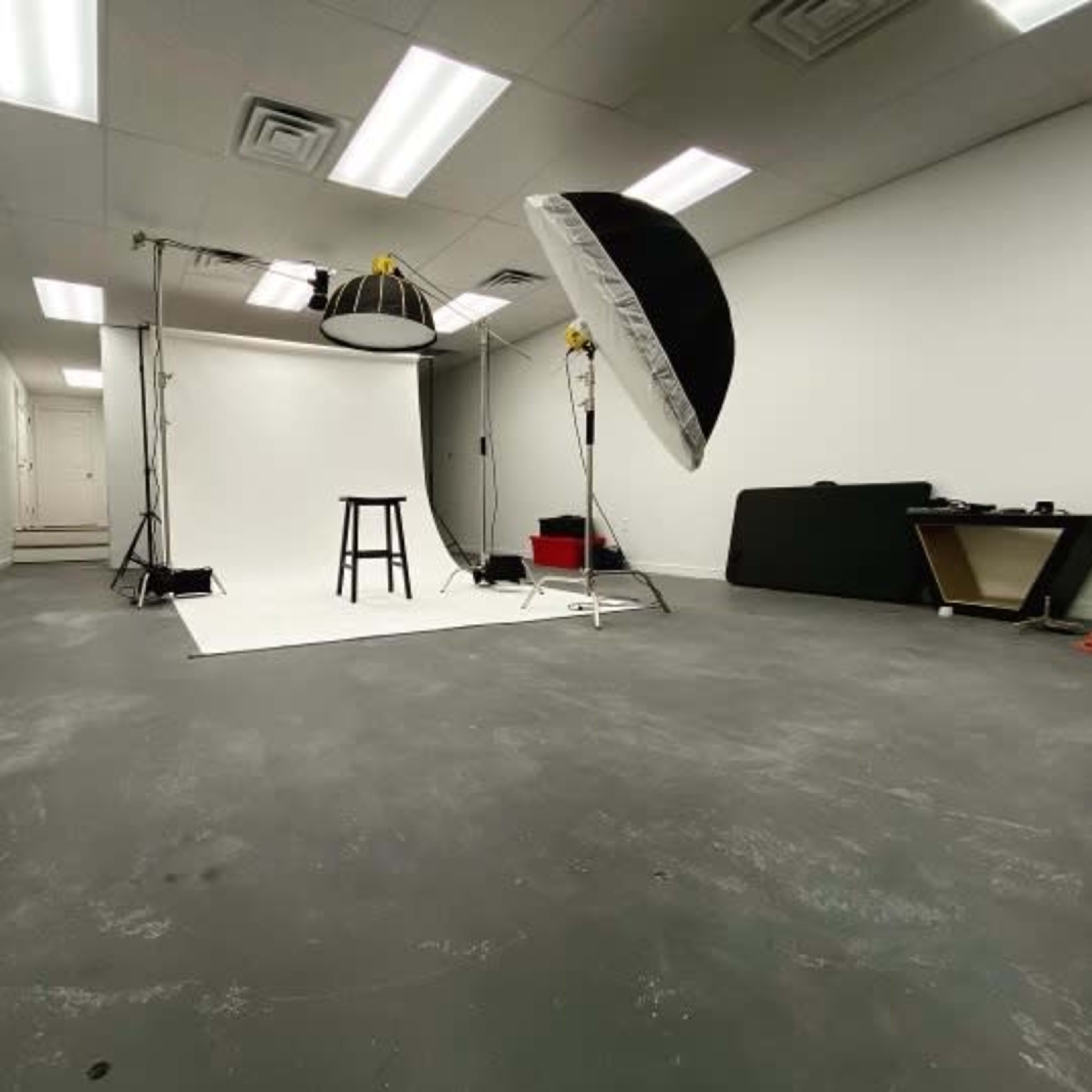 A photography studio with a white backdrop, lighting equipment, and a stool set up in an empty room.
