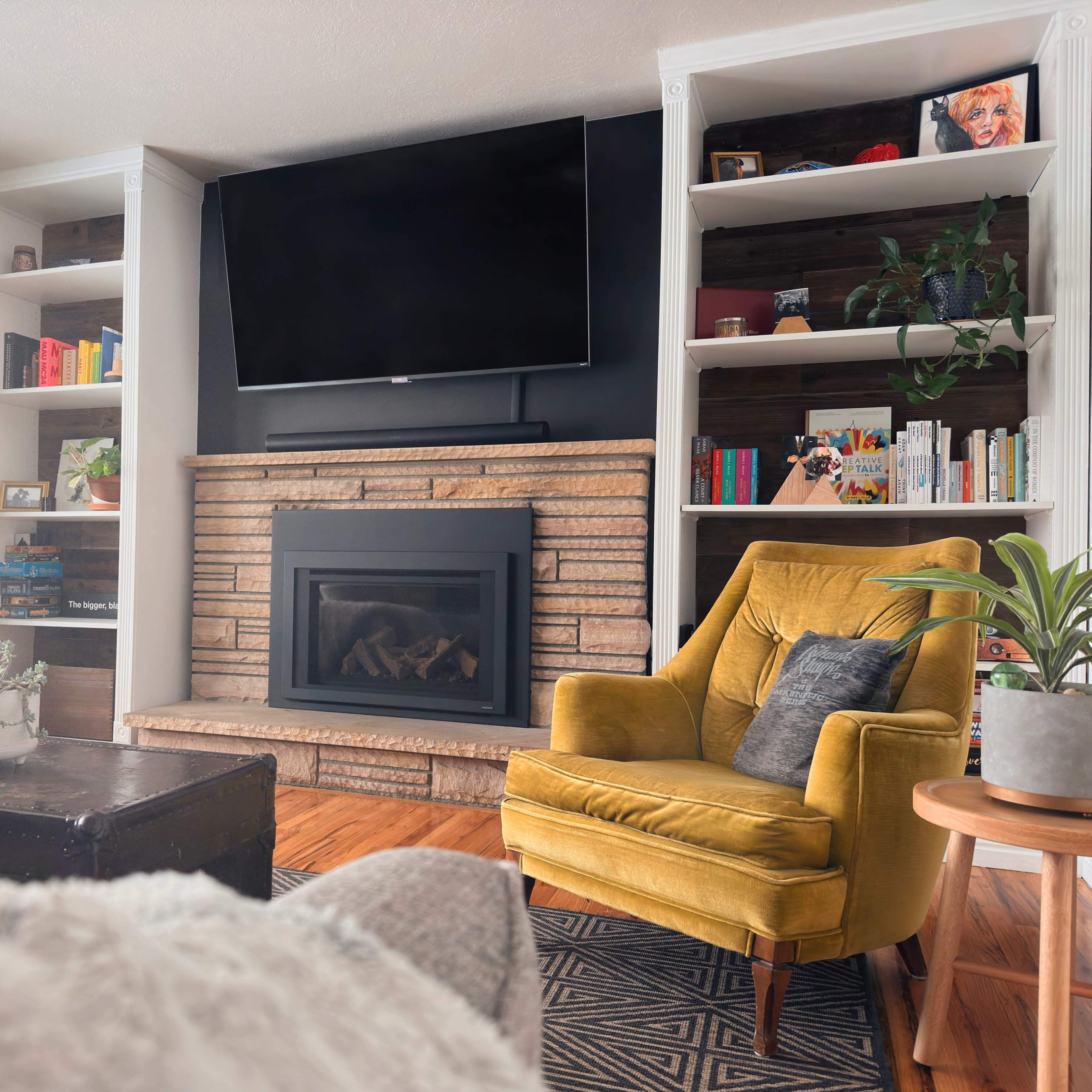 A cozy living room featuring a stone fireplace, a mounted TV, a yellow armchair, and built-in bookshelves filled with books and decorative items.