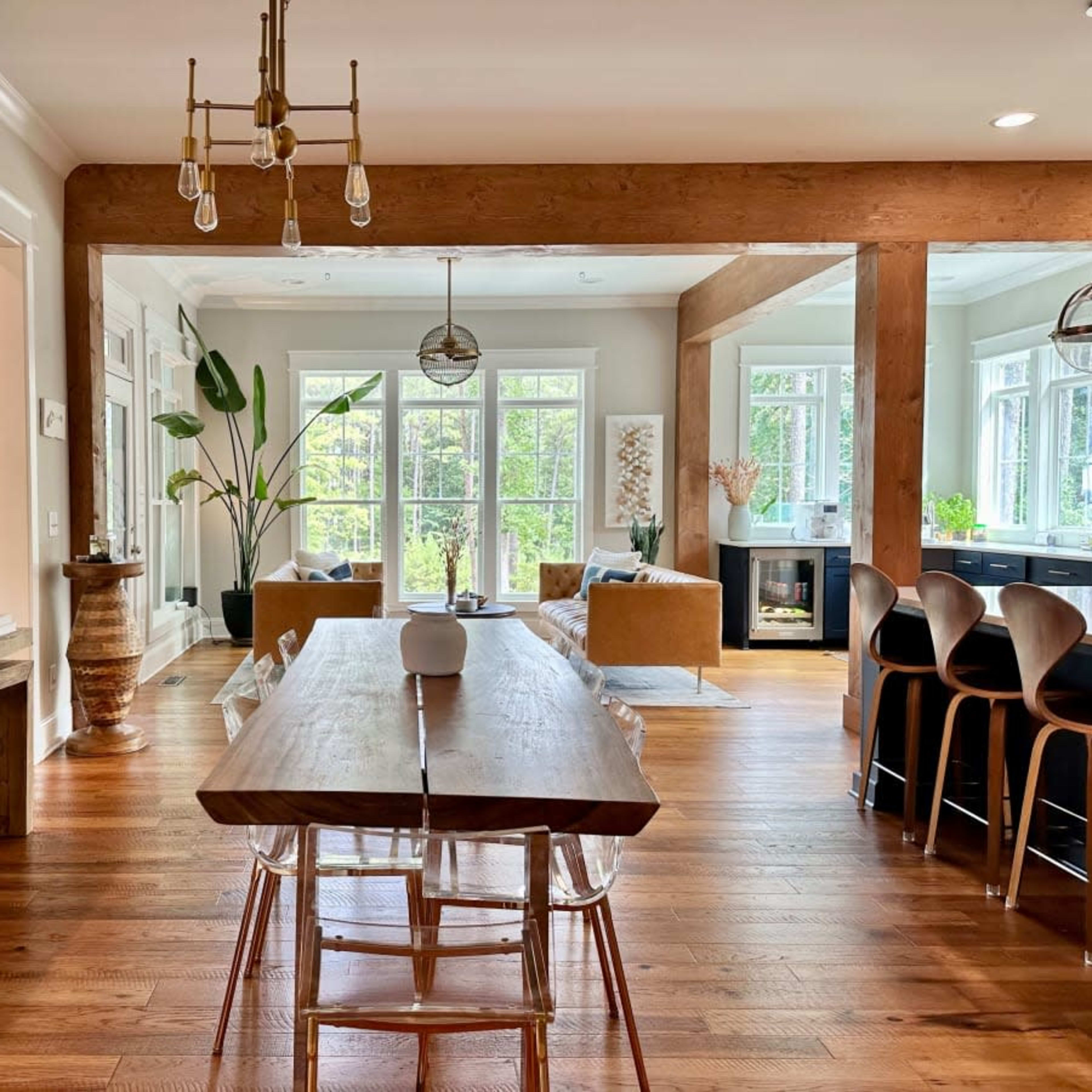 A spacious dining area features a large wooden table surrounded by clear chairs, with large windows and a view of greenery visible in the background.