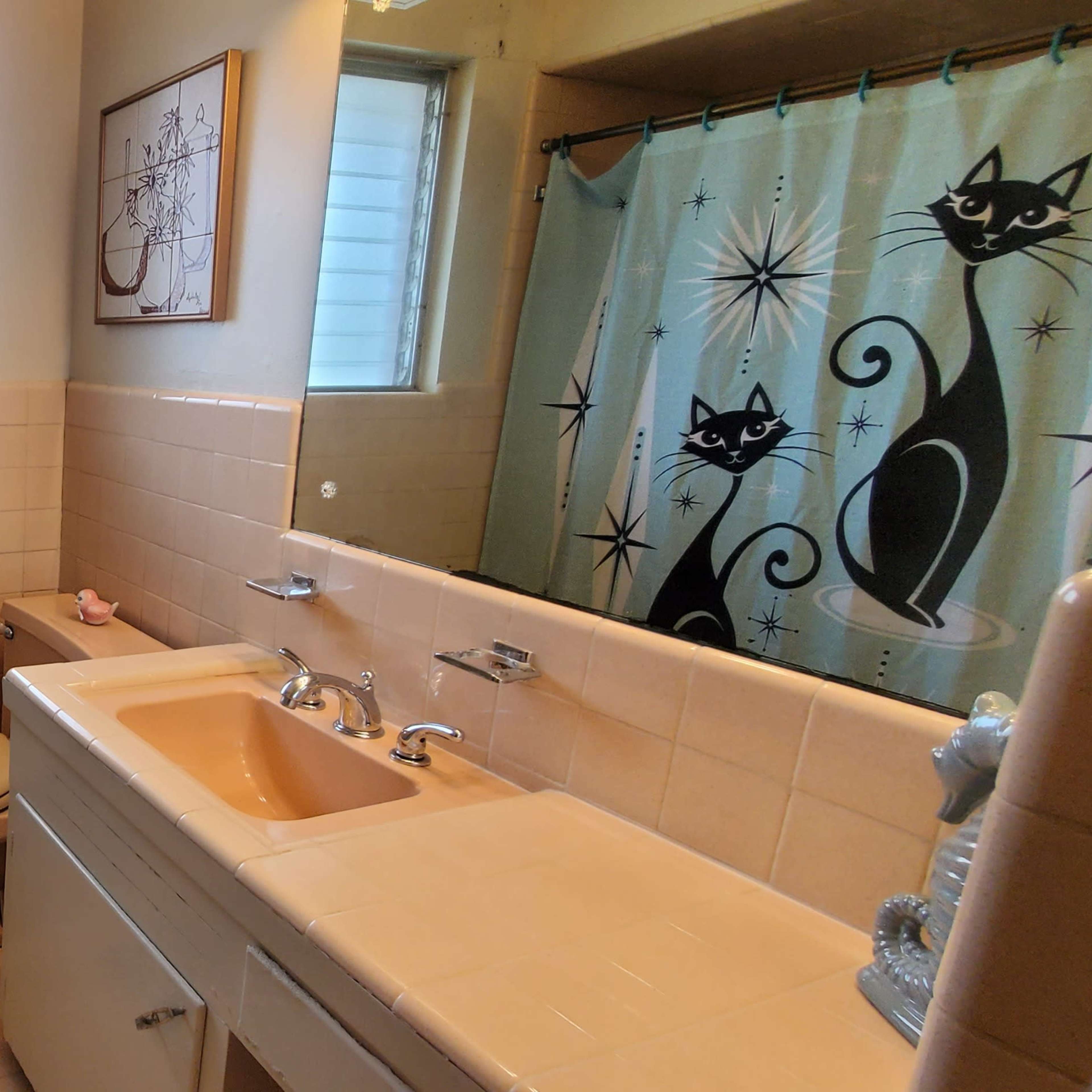 A bathroom with a pastel color scheme, featuring a toilet, sink with faucets, and a shower curtain adorned with black cat designs.