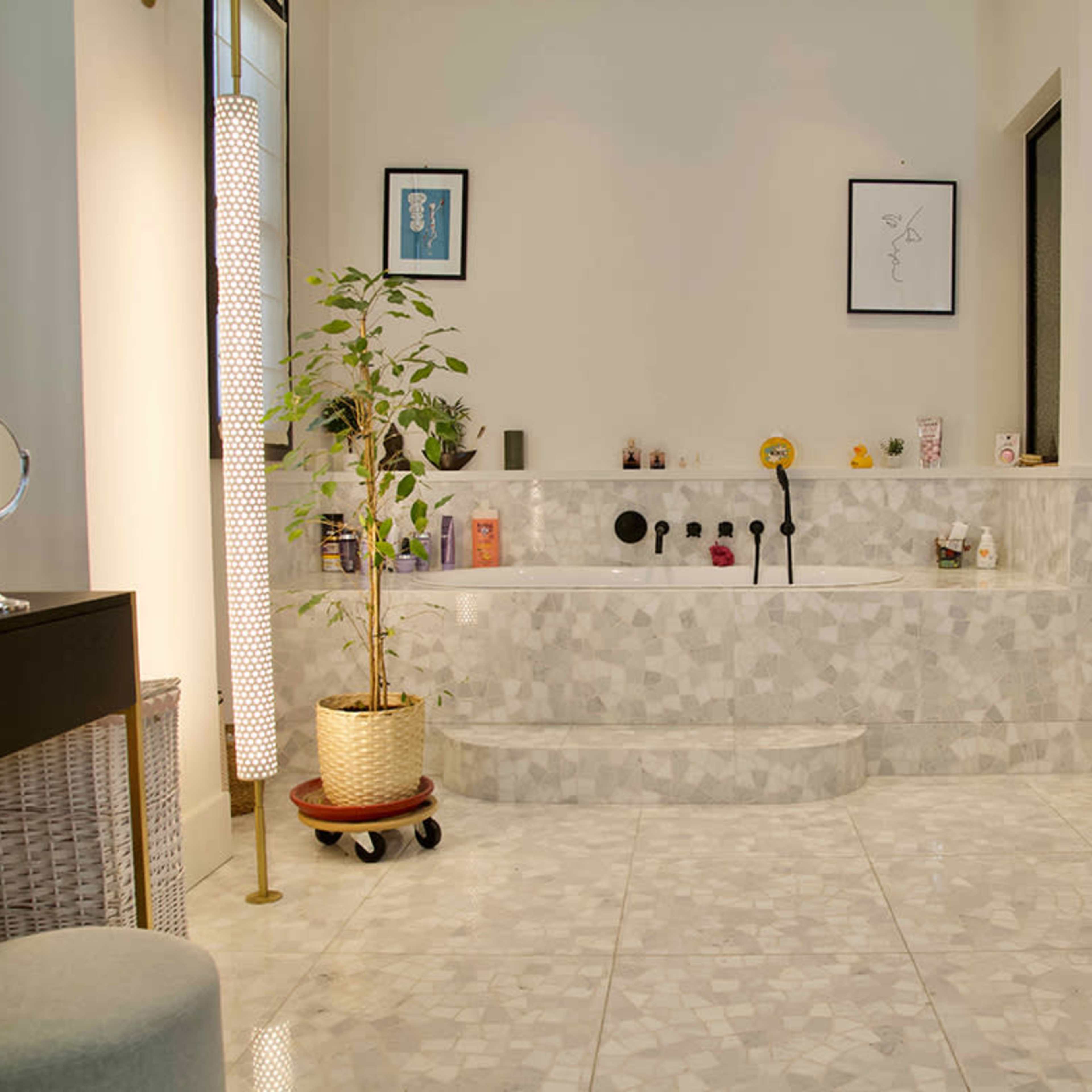 A modern bathroom featuring a marble bathtub, a small potted plant, framed art on the walls, and a textured floor with a shower area visible in the background.