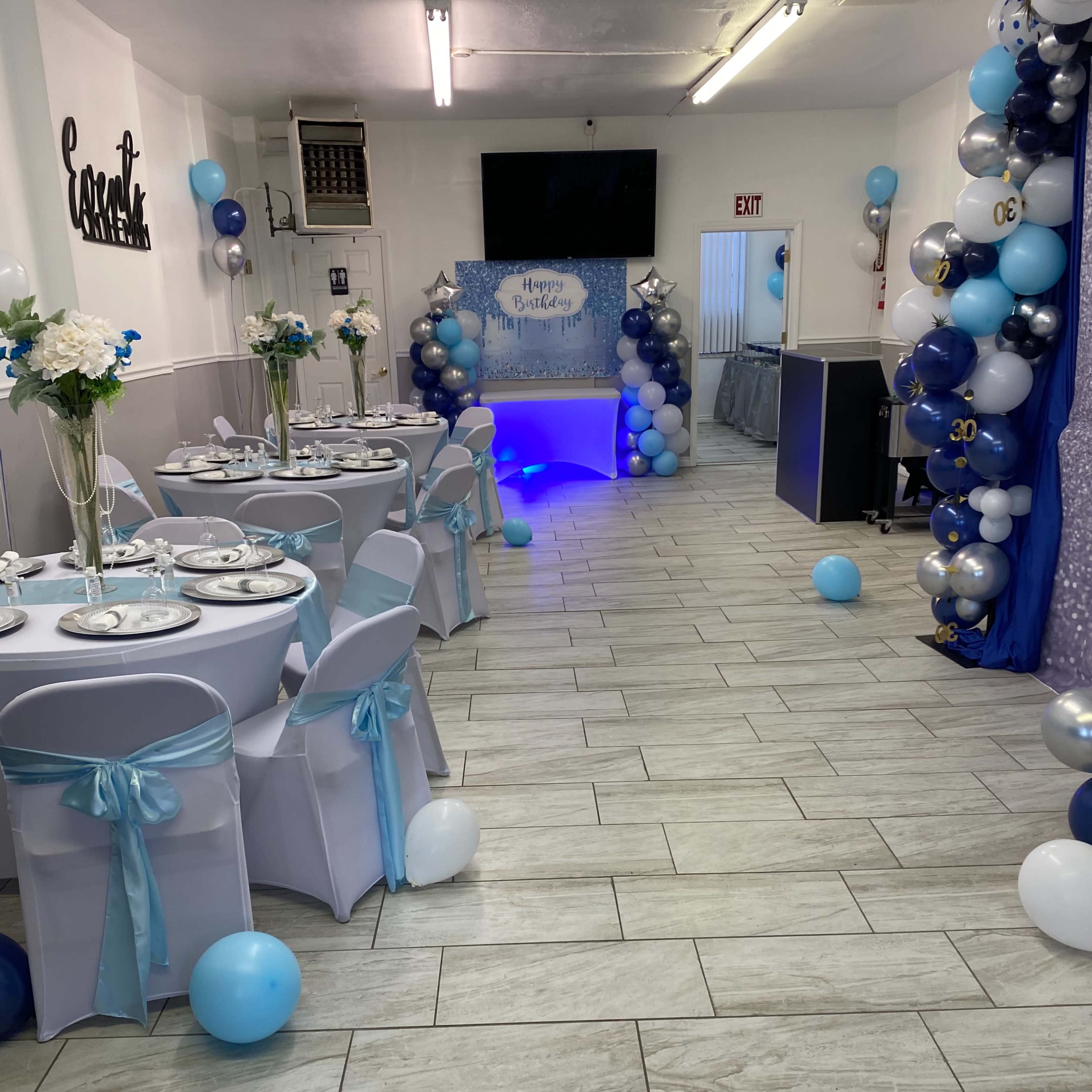 A decorated party space featuring tables with white tablecloths, blue and silver balloons, and a themed backdrop for a celebration.