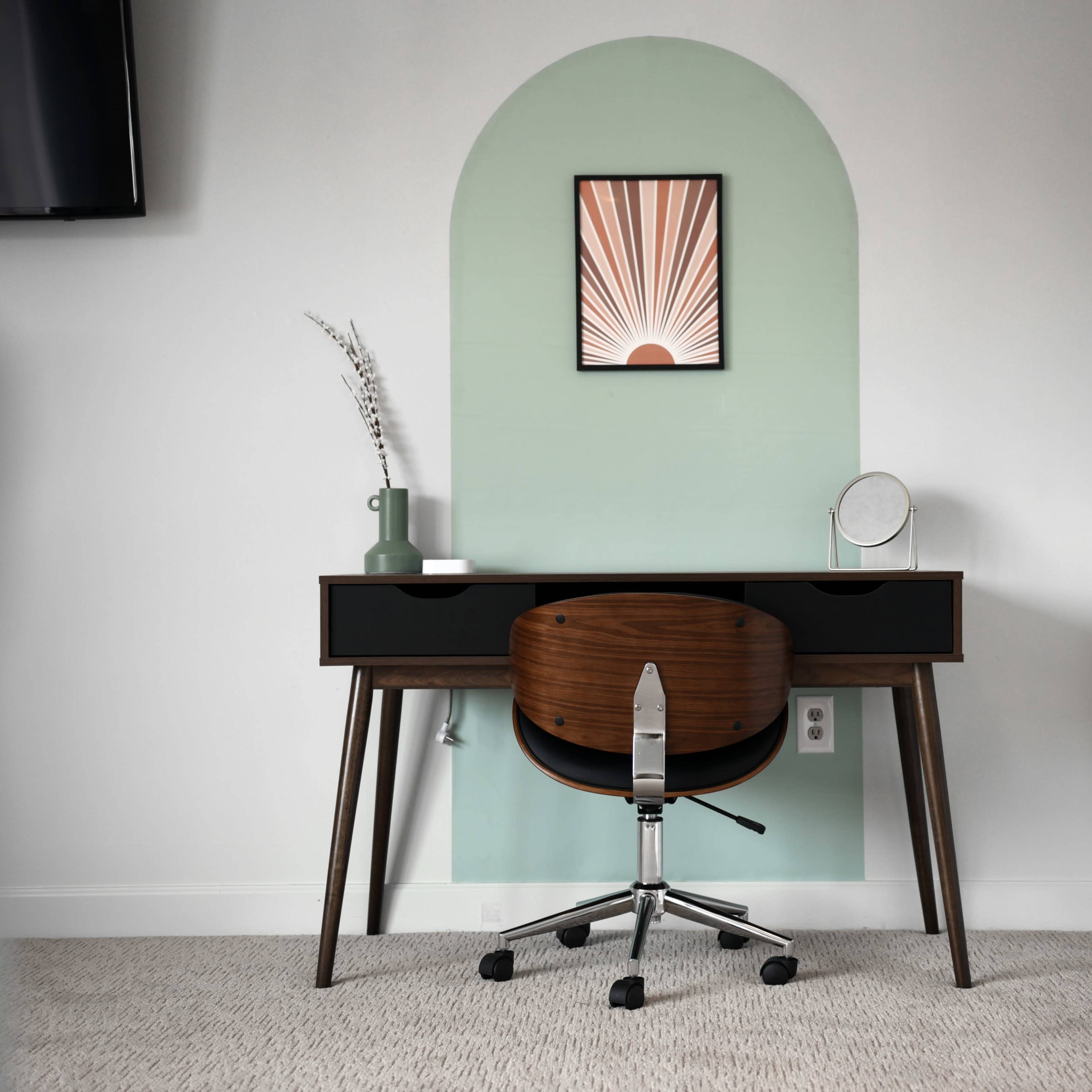 A minimalist desk with a chair is positioned against a light green wall featuring an arched design and a framed artwork.