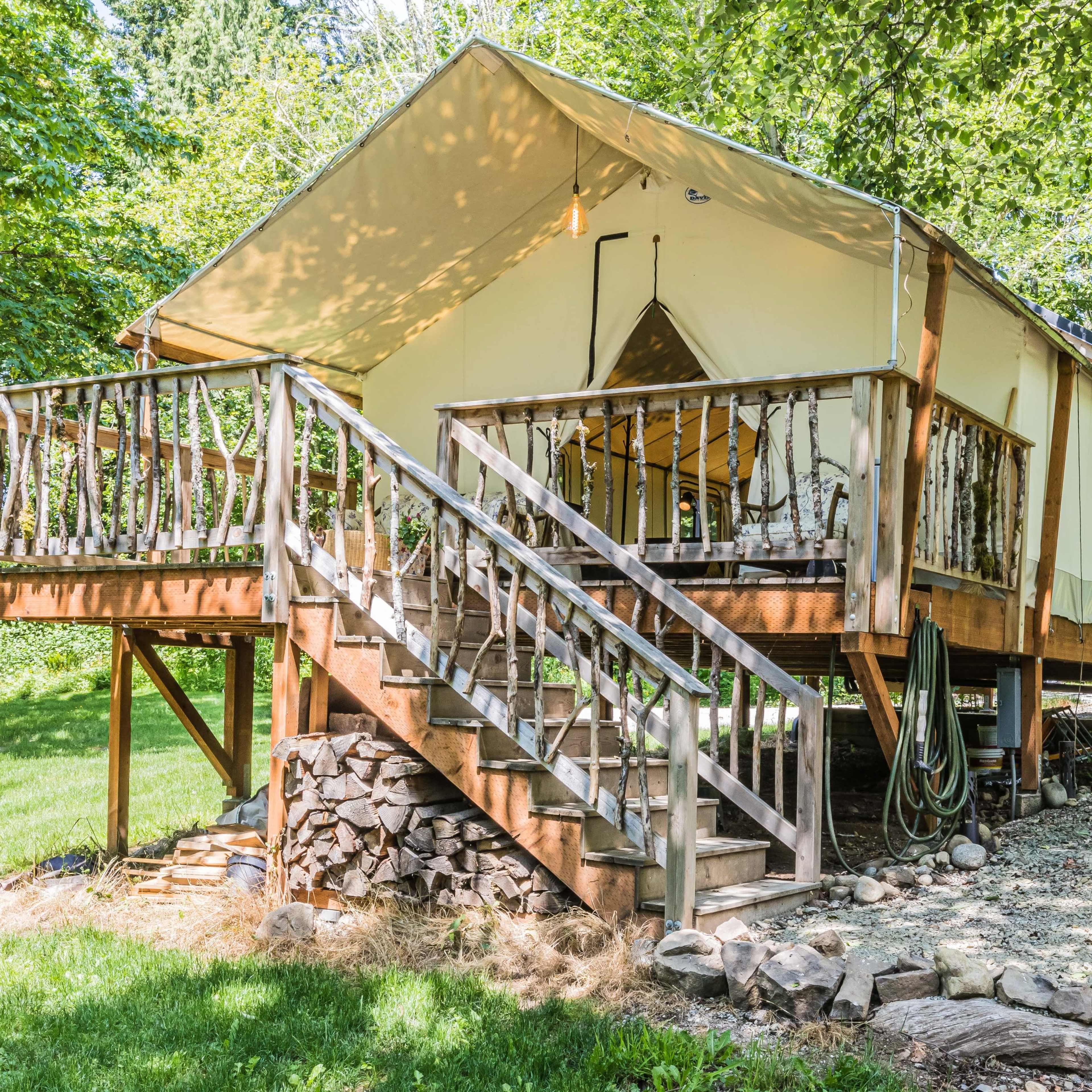A tent cabin sits elevated on a wooden platform with a set of stairs leading up to a deck surrounded by trees.