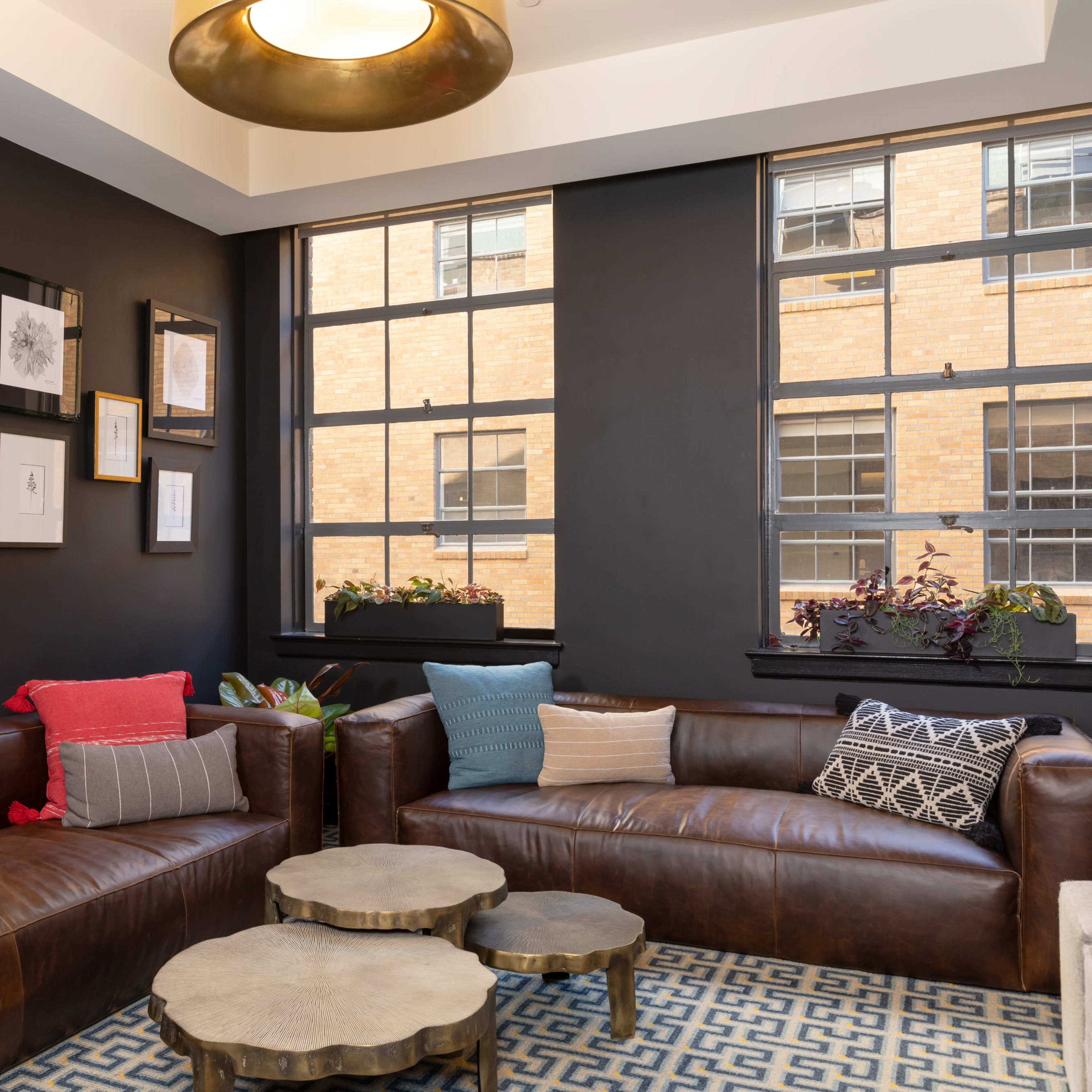 A cozy seating area features two leather sofas and a set of metallic coffee tables, illuminated by natural light streaming through large windows adorned with potted plants.