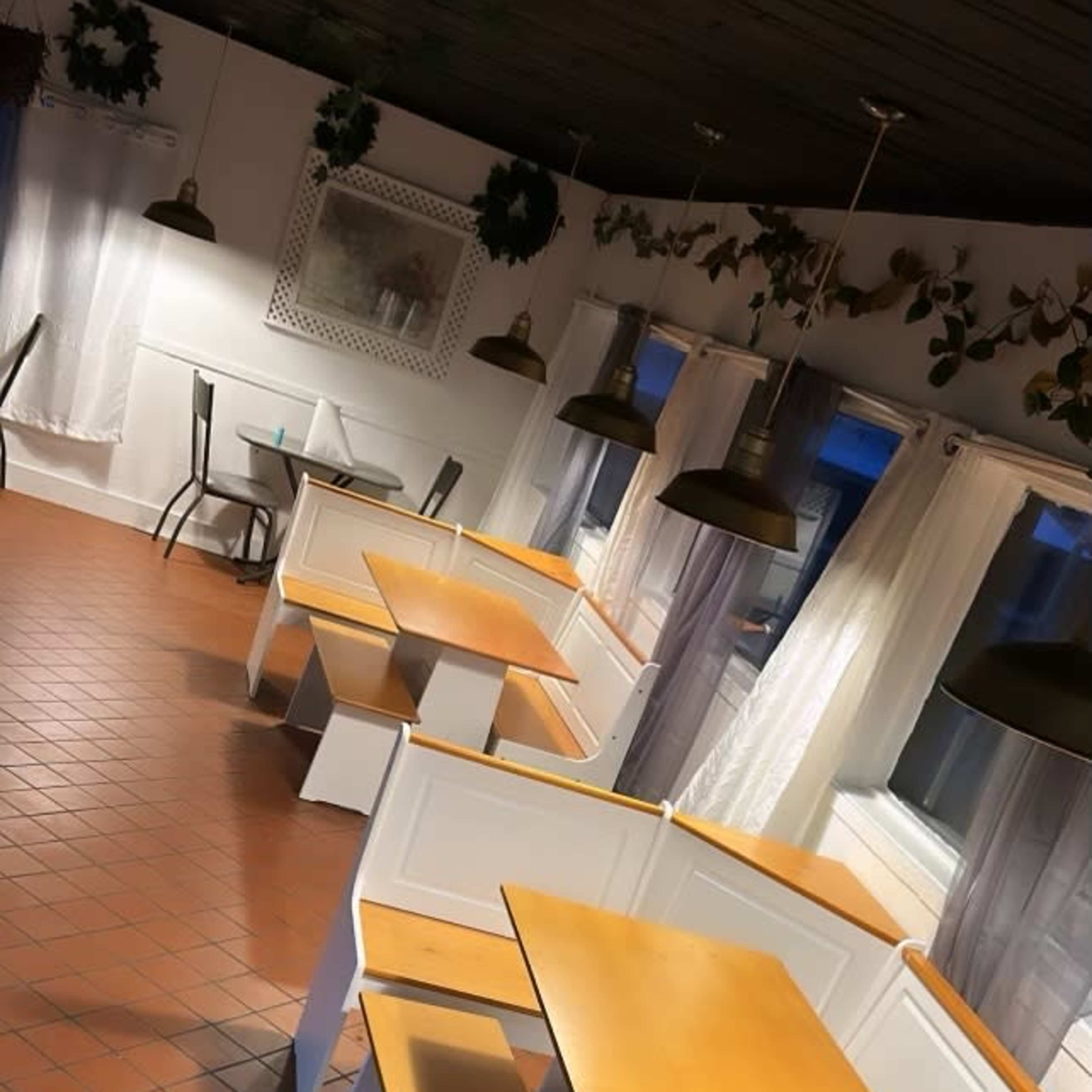 A light-filled dining area featuring multiple wooden tables and benches arranged in a square configuration, with large windows and hanging pendant lights.