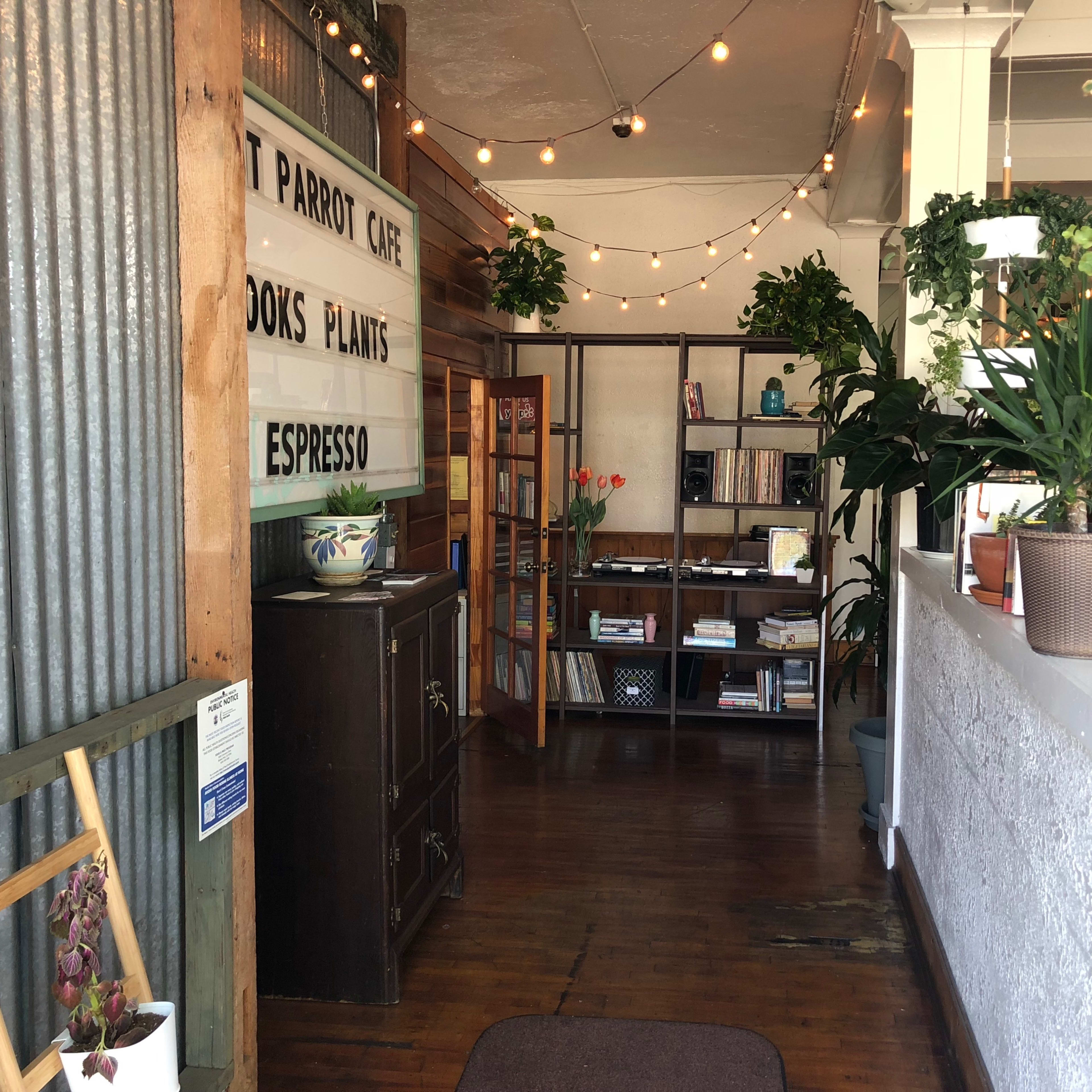 A cozy interior of a café featuring a wooden floor, a bookshelf, potted plants, and a lighted marquee sign above the entrance.