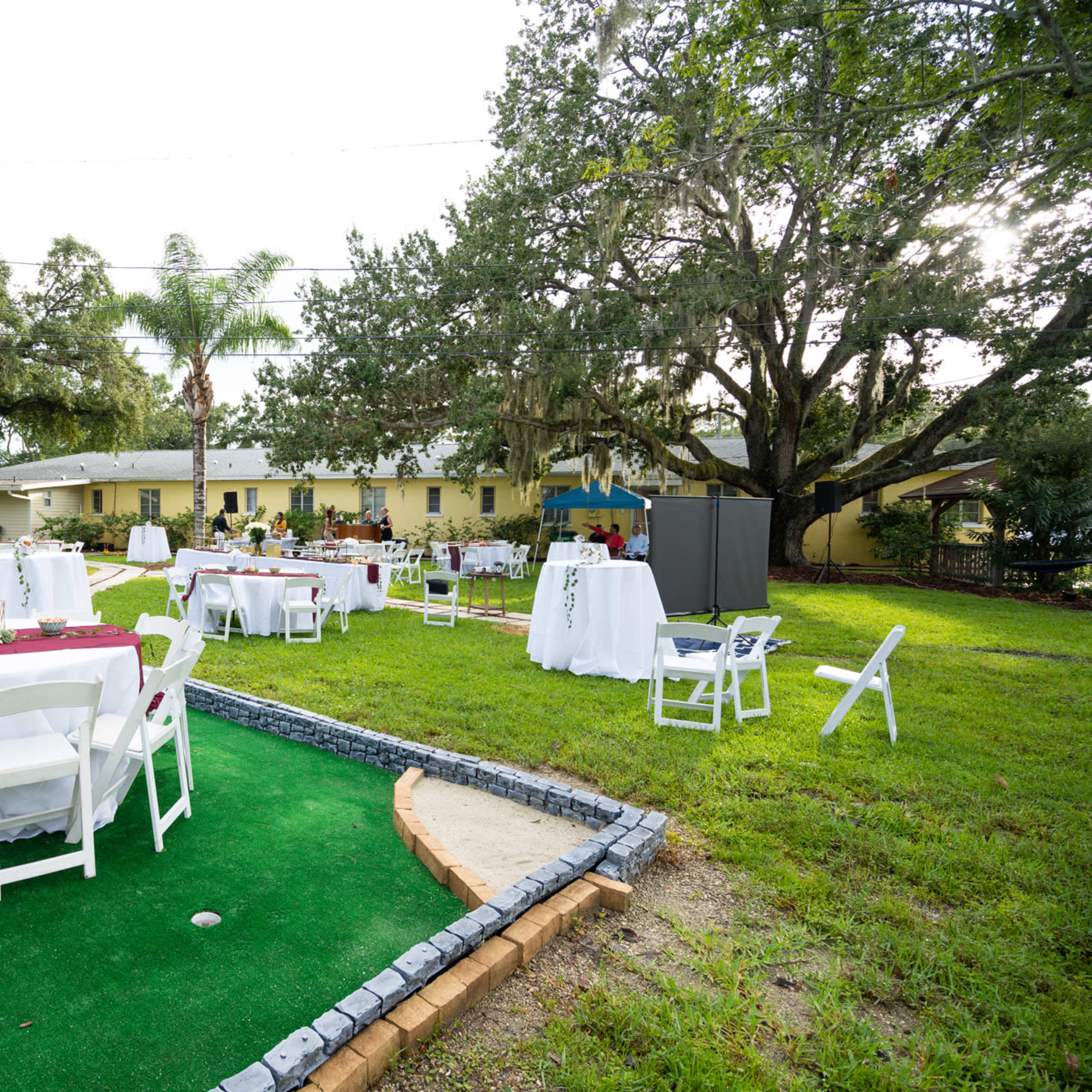 A spacious outdoor area is set up for an event, featuring several round tables covered with white tablecloths and maroon accents placed on green turf.