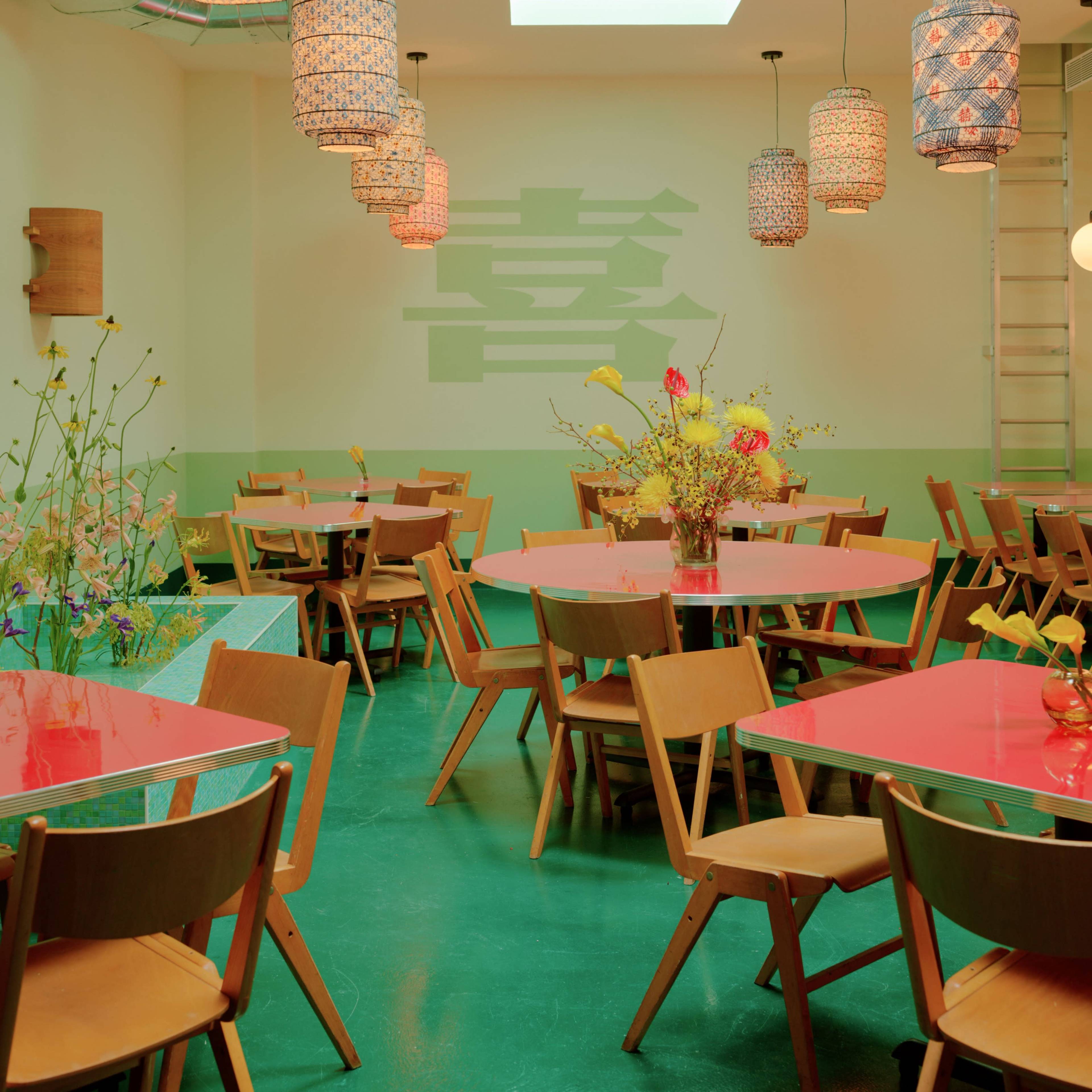 A brightly lit restaurant interior featuring wooden chairs and tables with colorful tabletops, adorned with hanging pendant lights and floral arrangements.