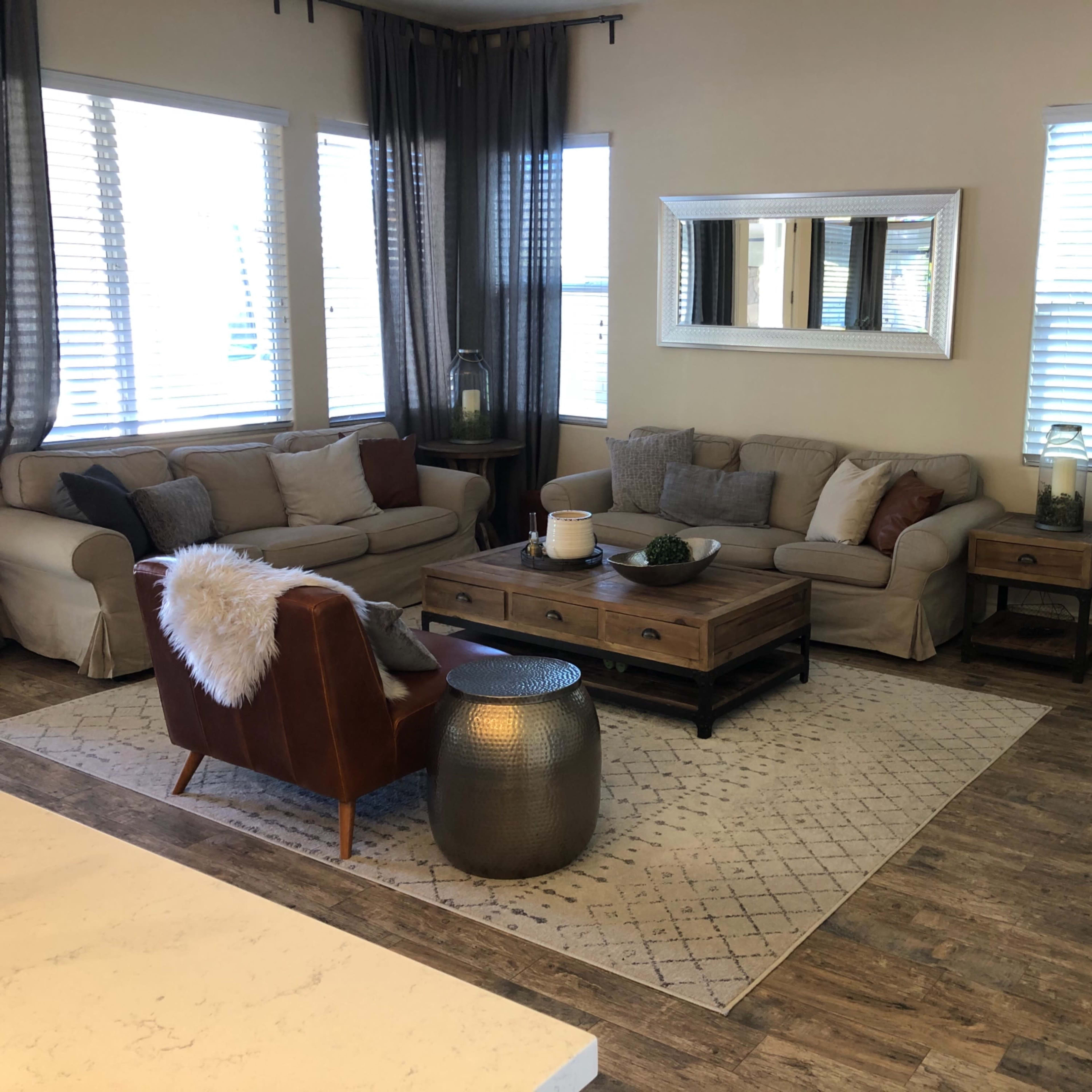 A living room featuring a sectional sofa, a coffee table, a plant, and large windows with sheer curtains.