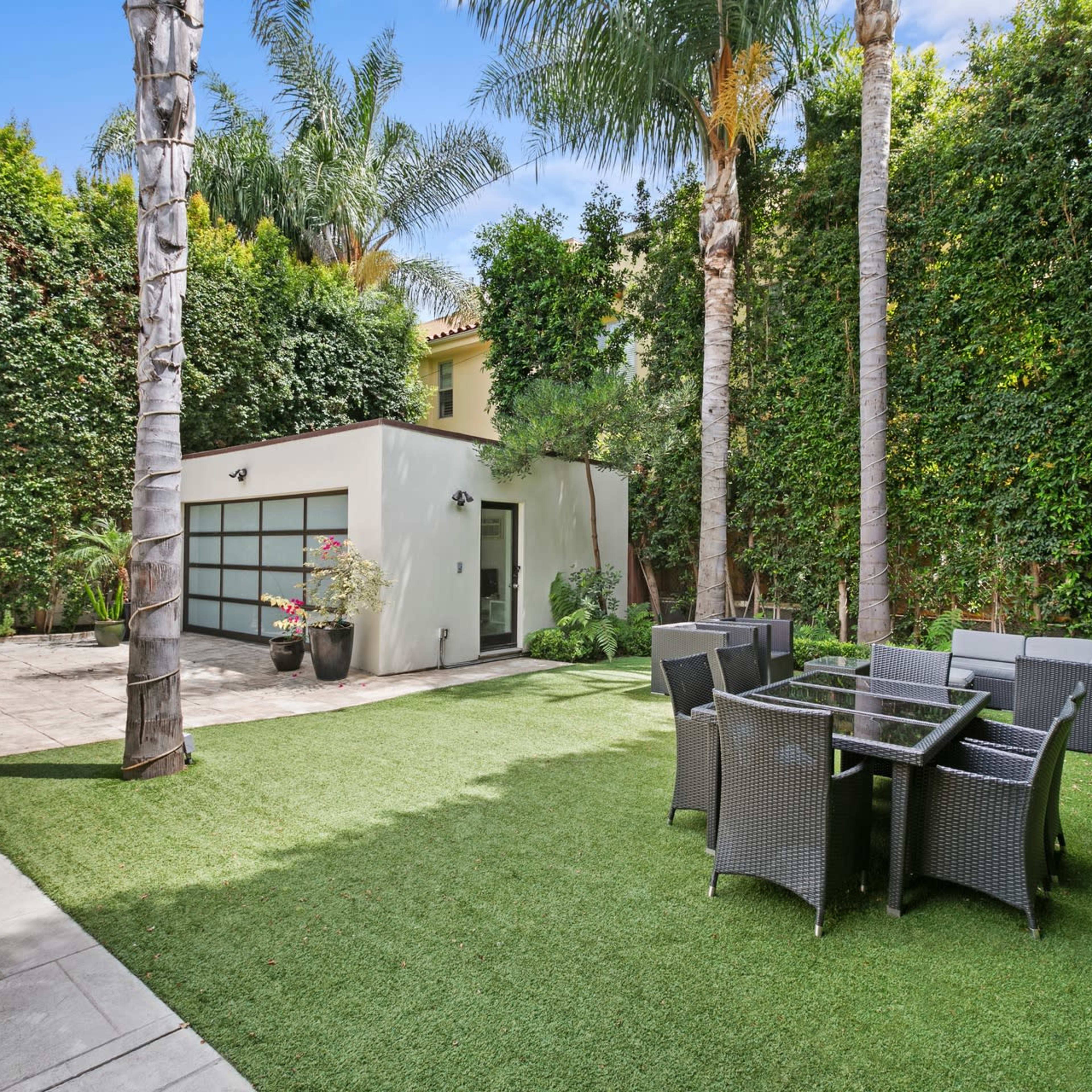 A landscaped outdoor area features a small building, a grassy lawn, and a dining set surrounded by tall palm trees and vibrant greenery.
