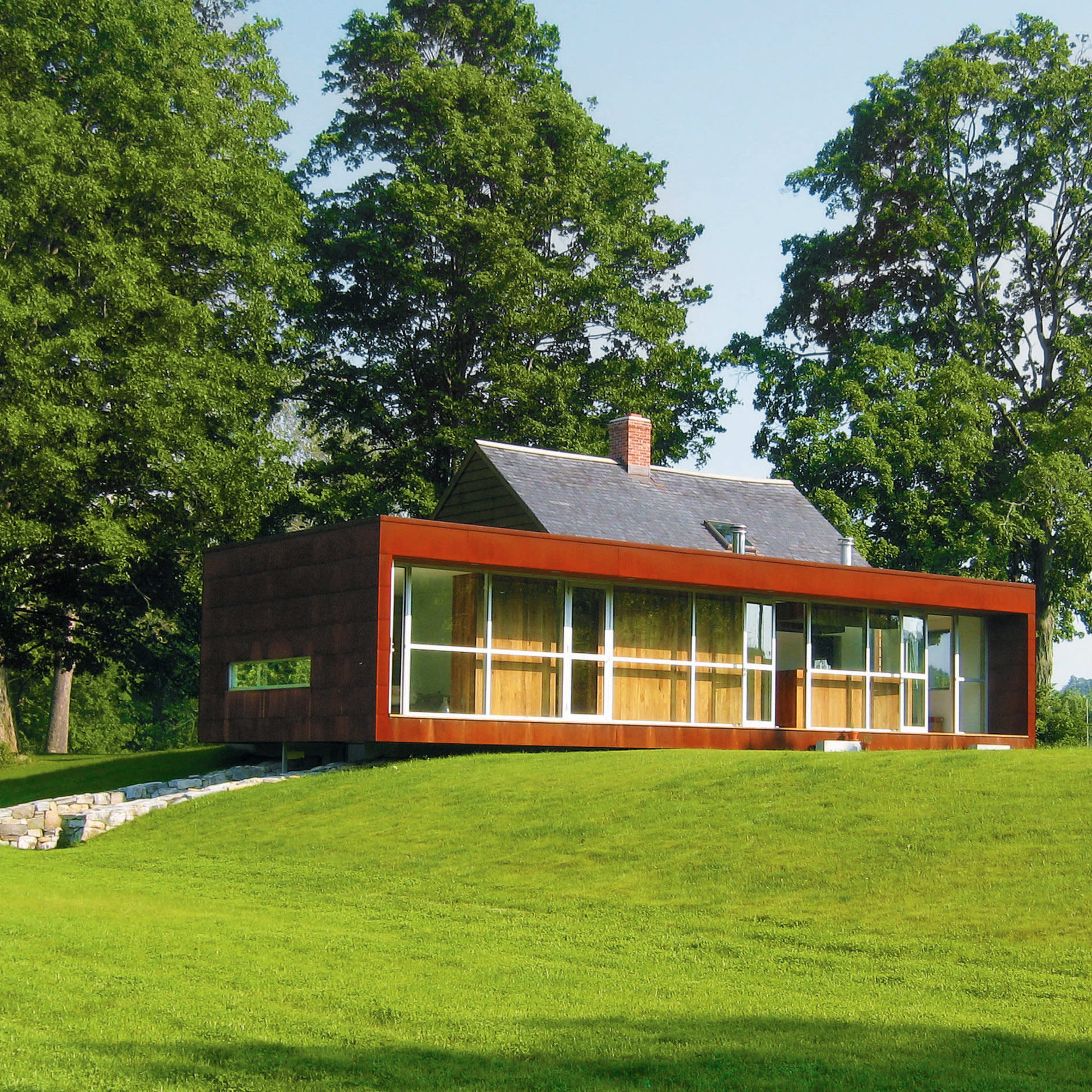 A modern house with a combination of wooden and glass elements sits on a grassy slope, surrounded by large trees.