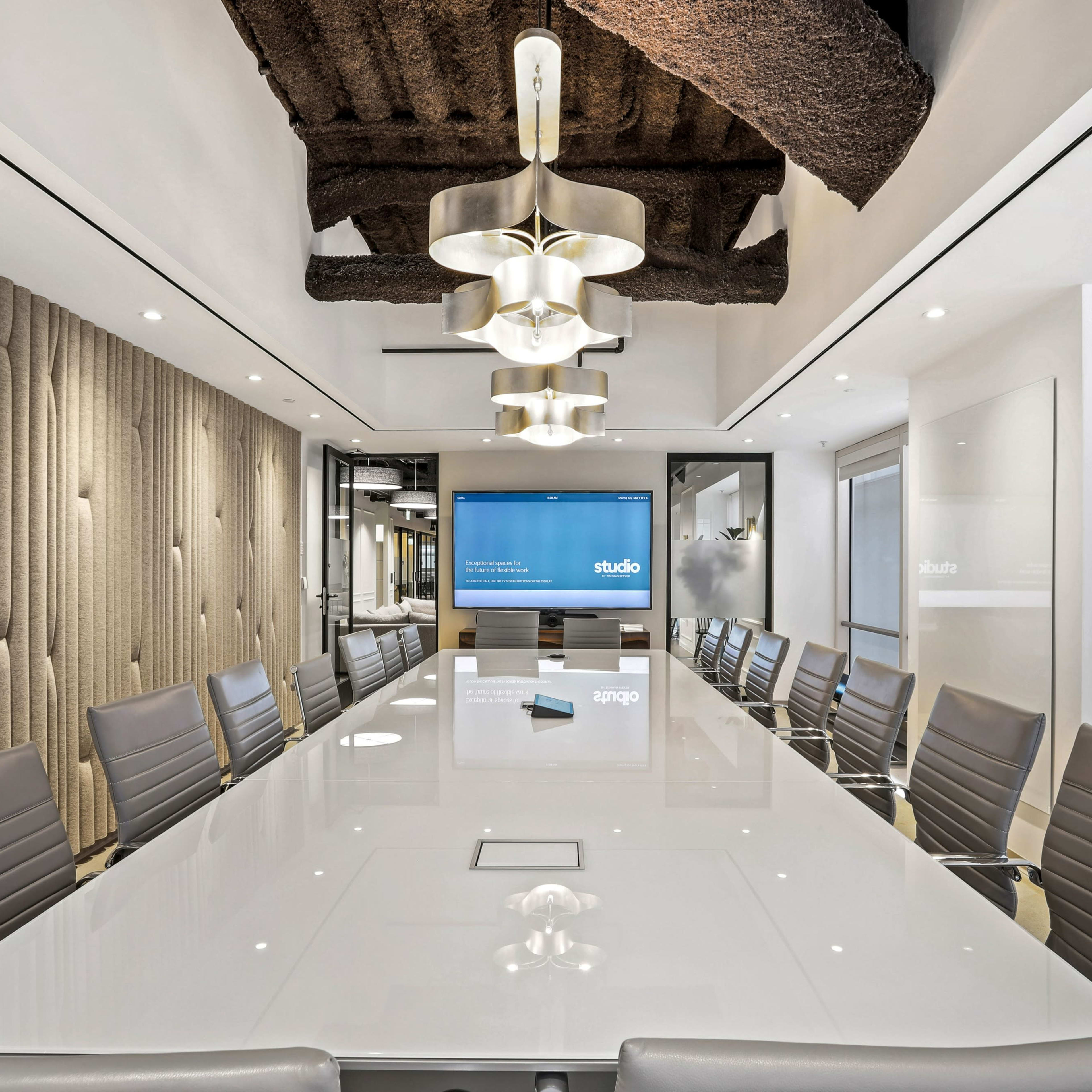 A modern conference room with a large white table surrounded by gray chairs, featuring a screen on one wall and decorative ceiling beams.