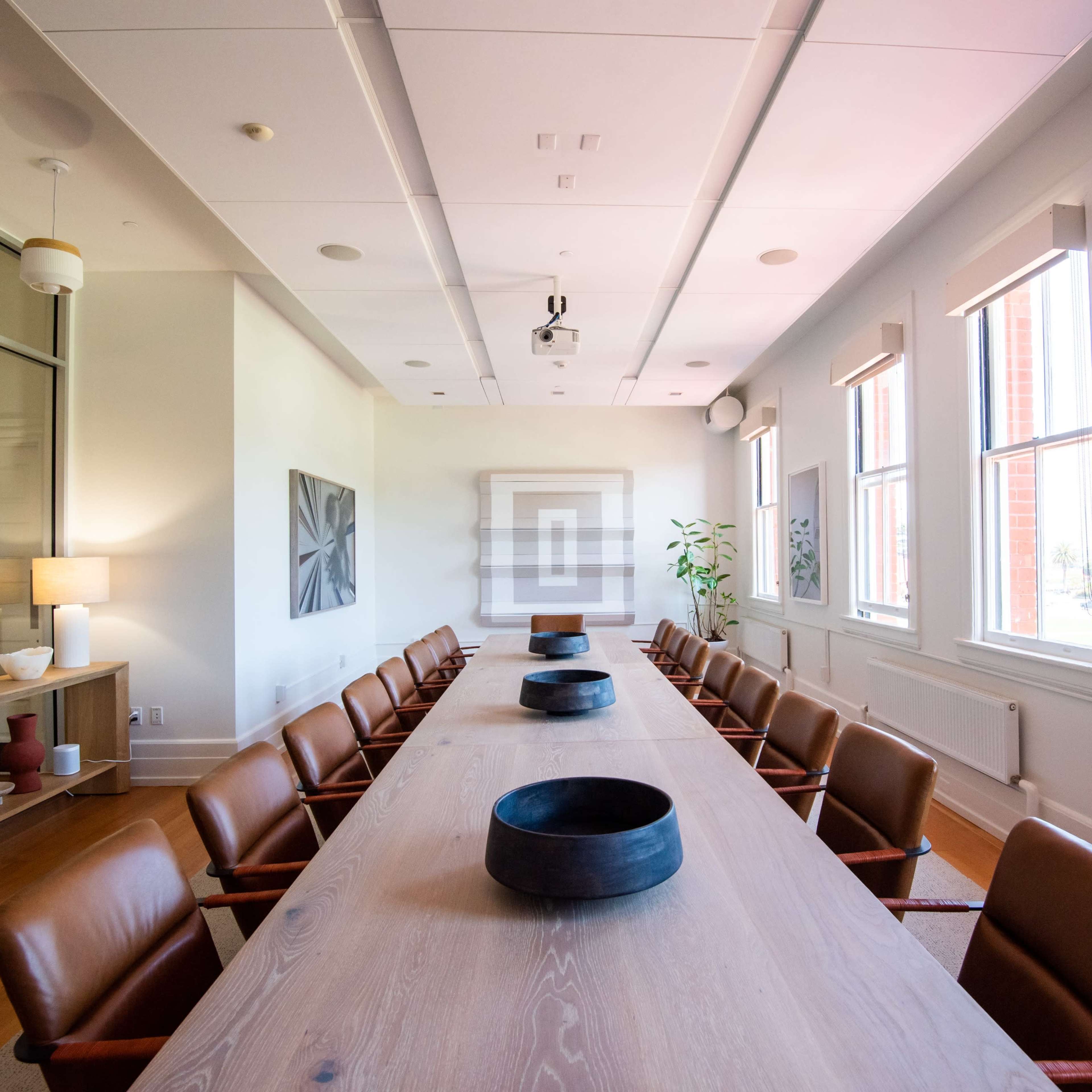 A long wooden conference table surrounded by leather chairs is set in a brightly lit meeting room with large windows and modern decor.