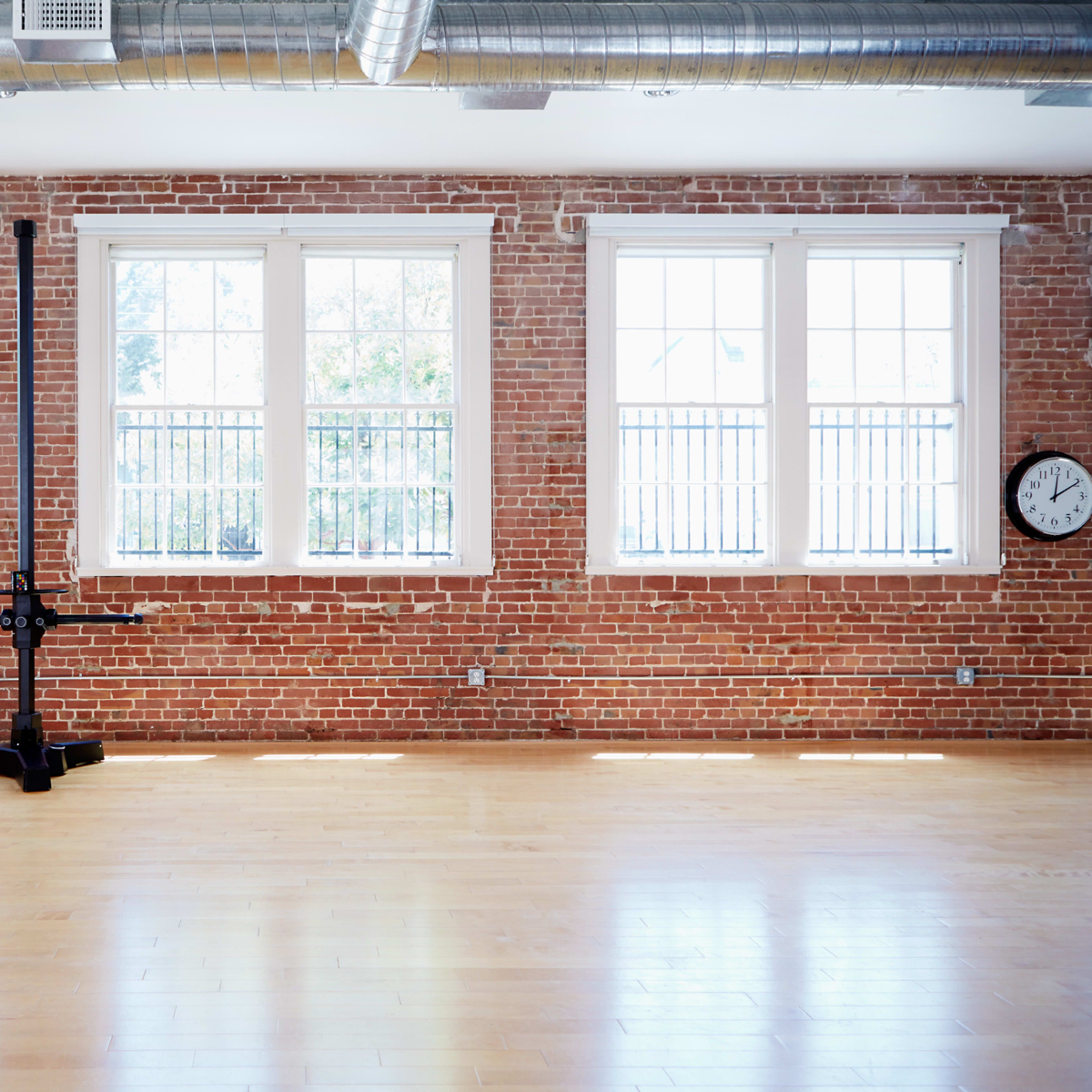 A spacious room with exposed brick walls, large windows, and a wooden floor, featuring a clock and an exercise equipment stand in the center.
