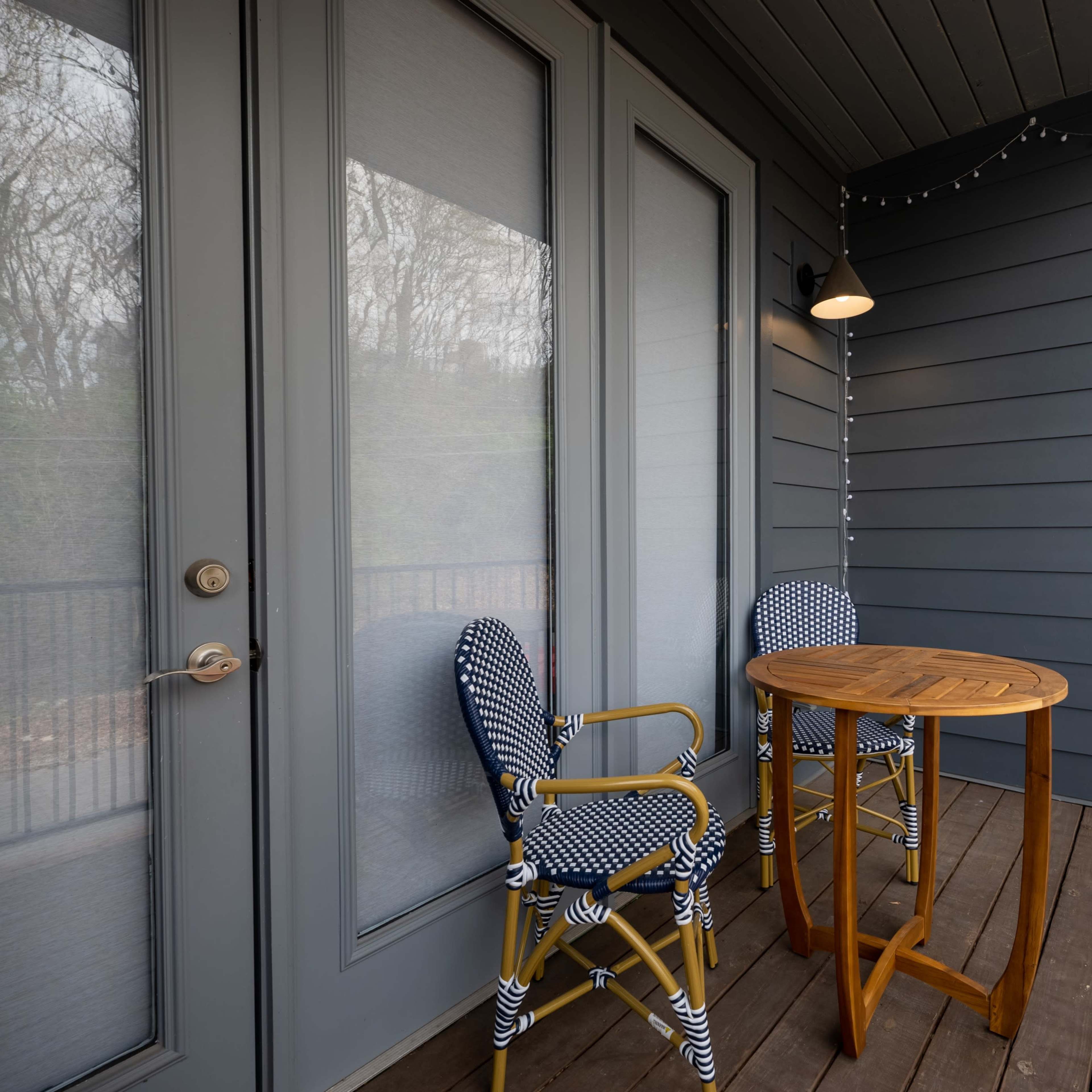A small outdoor balcony featuring a round wooden table and two blue and white striped chairs, with a light fixture and decorative string lights above.
