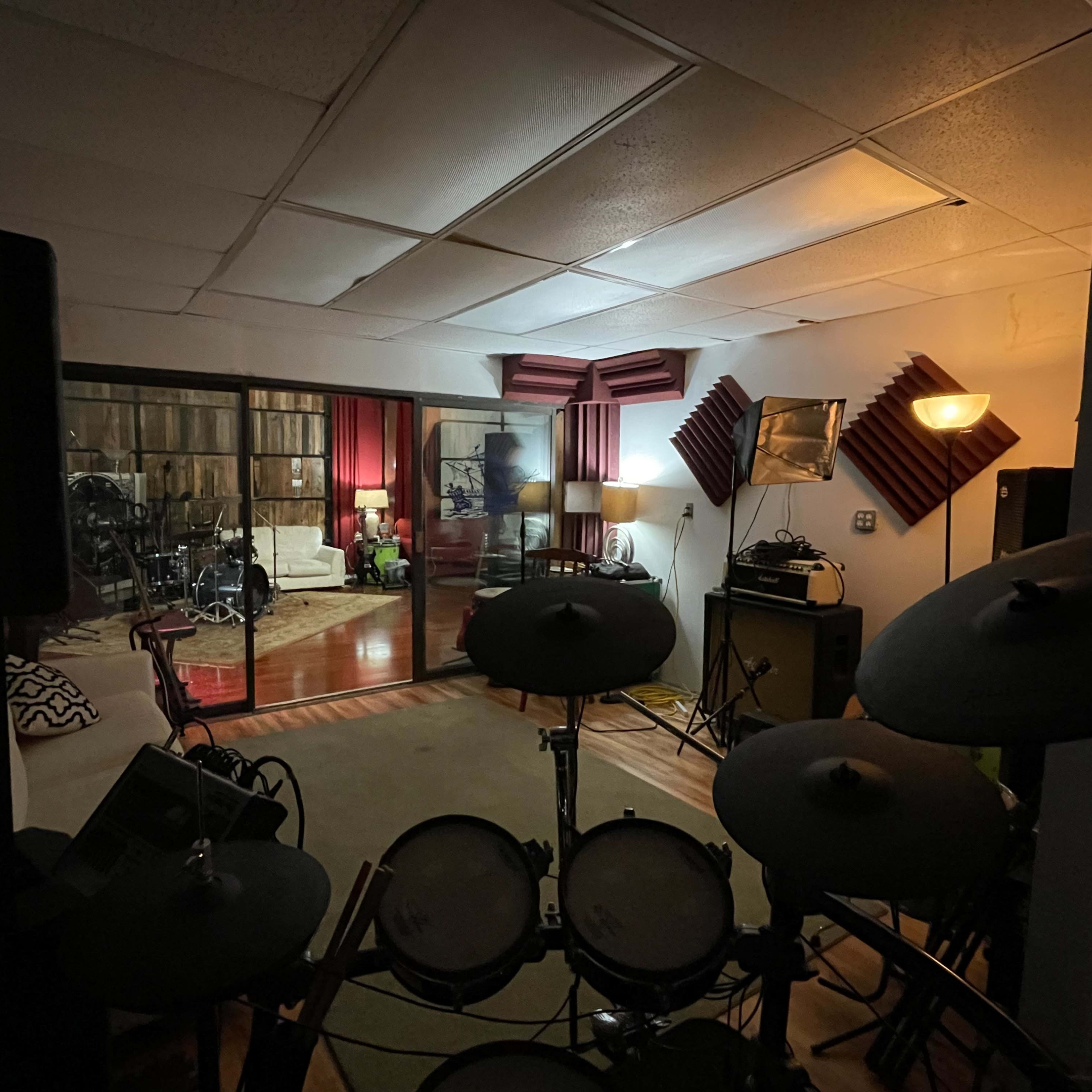 A dimly lit music studio with electronic drums in the foreground and a cozy lounge area featuring a couch and lamps in the background.