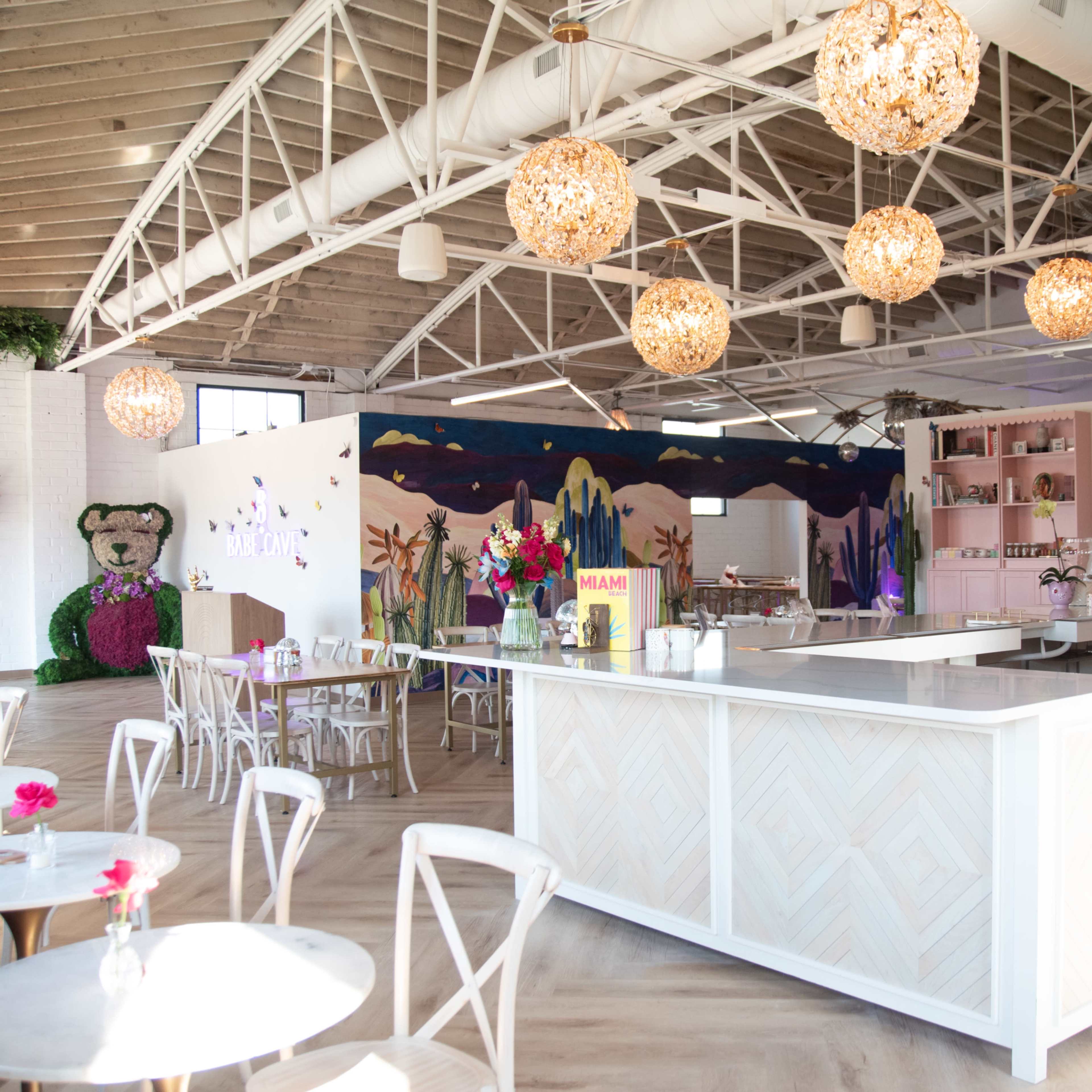 A brightly lit café with a white counter, round tables with chairs, and decorative pendant lights hanging from the ceiling.